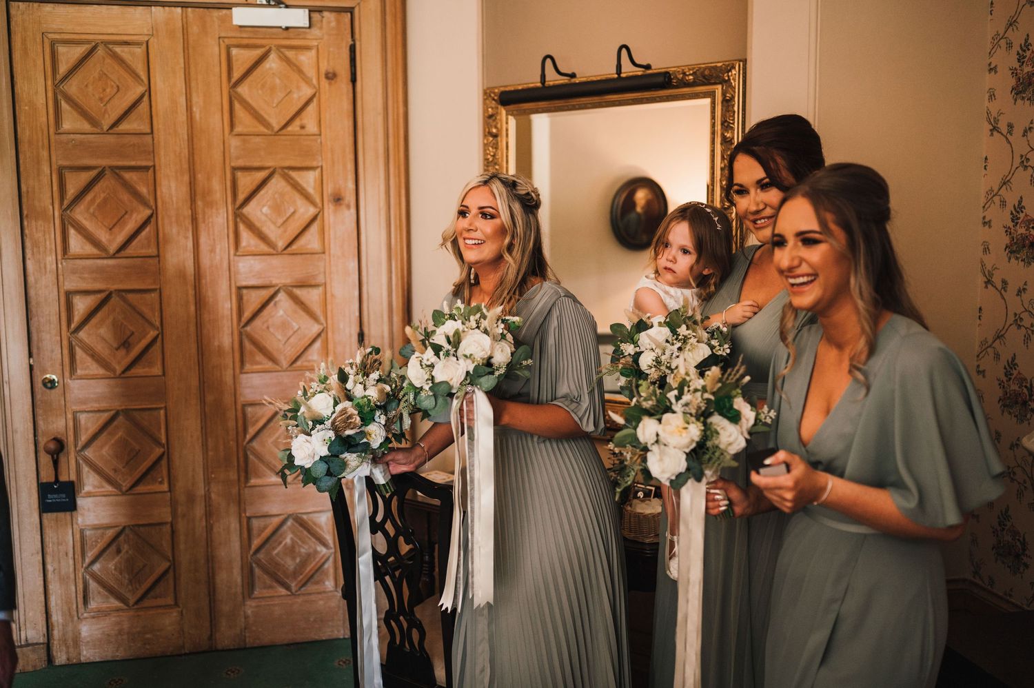 Bridesmaids in sage dresses hold white floral bouquets while standing in an elegant venue.