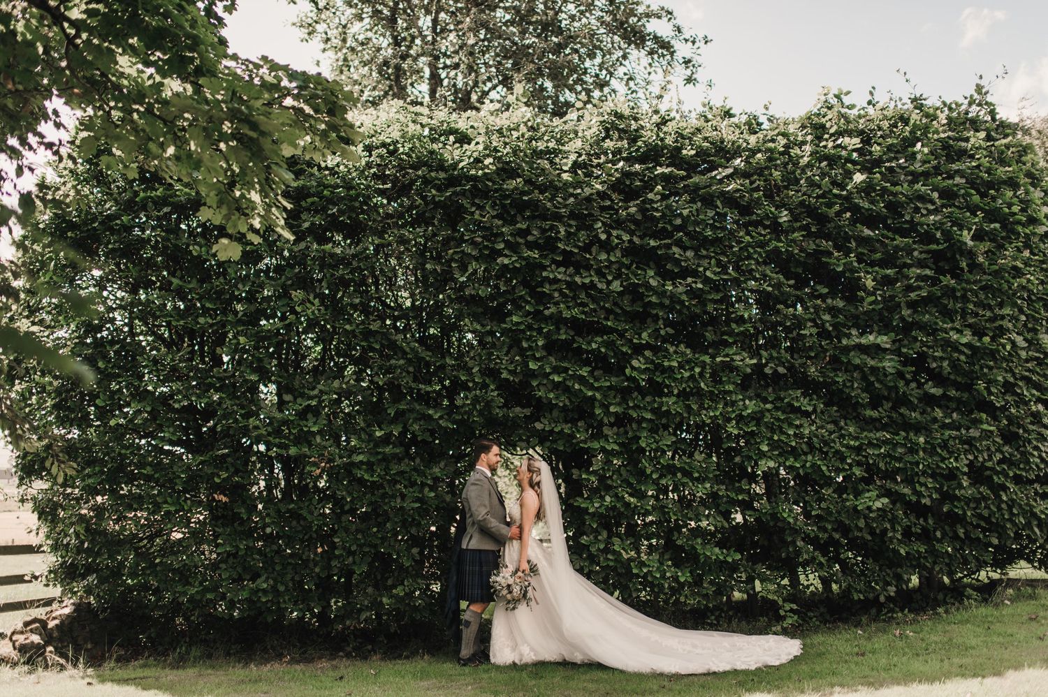 A wedding couple embraces in front of a tall hedge wall in a garden setting.