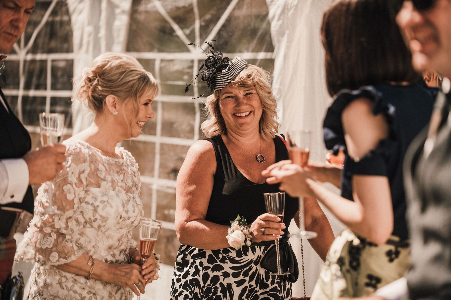 Wedding guests share laughs and drinks during an outdoor tent reception celebration.