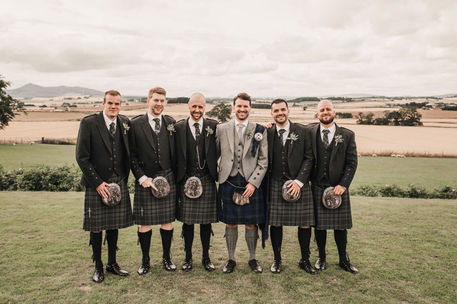 Wedding party in traditional Scottish kilts posed for formal photo on grassy hillside.
