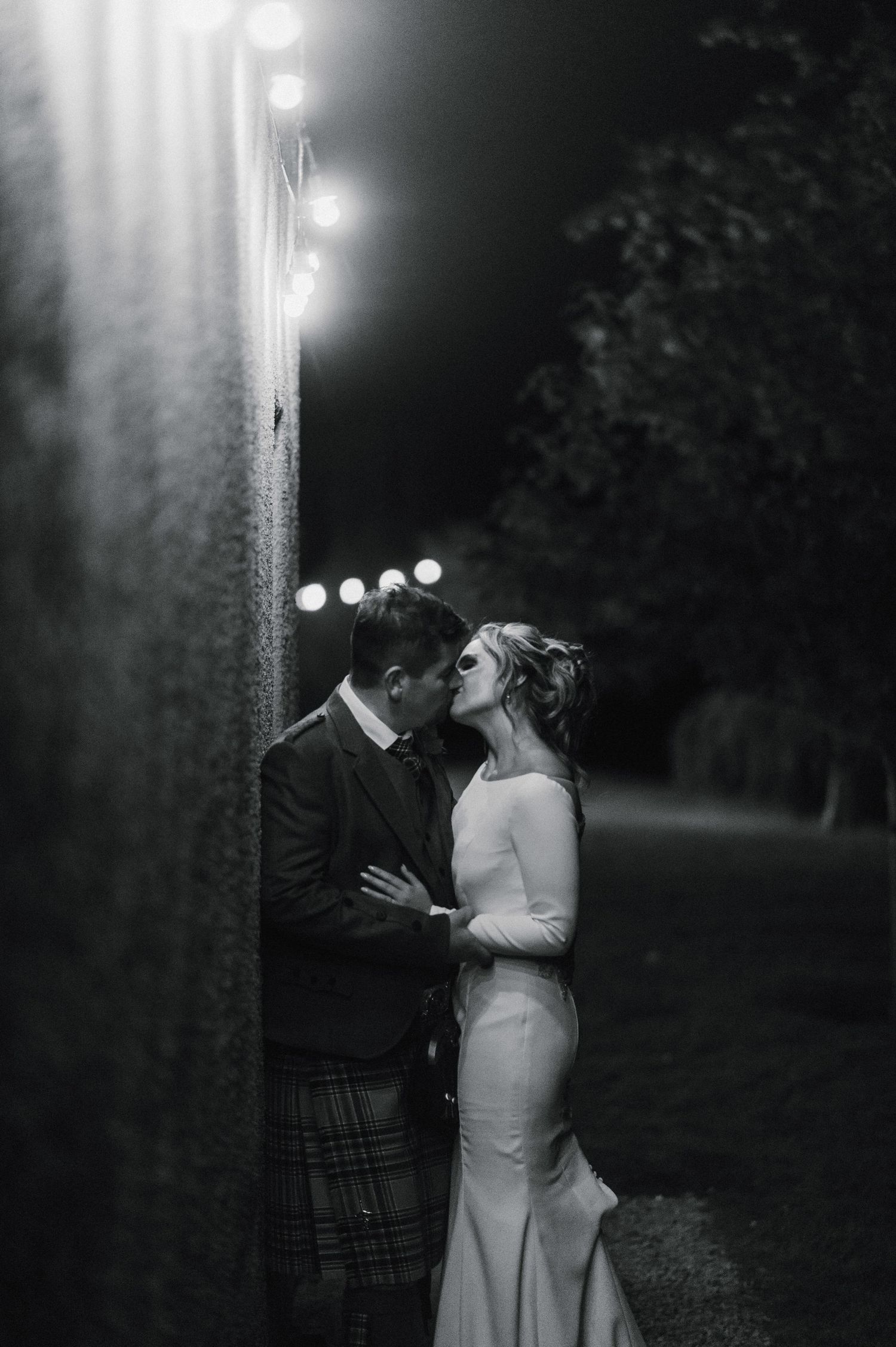 An intimate black and white portrait of newlyweds sharing a tender moment against a stone wall at night.