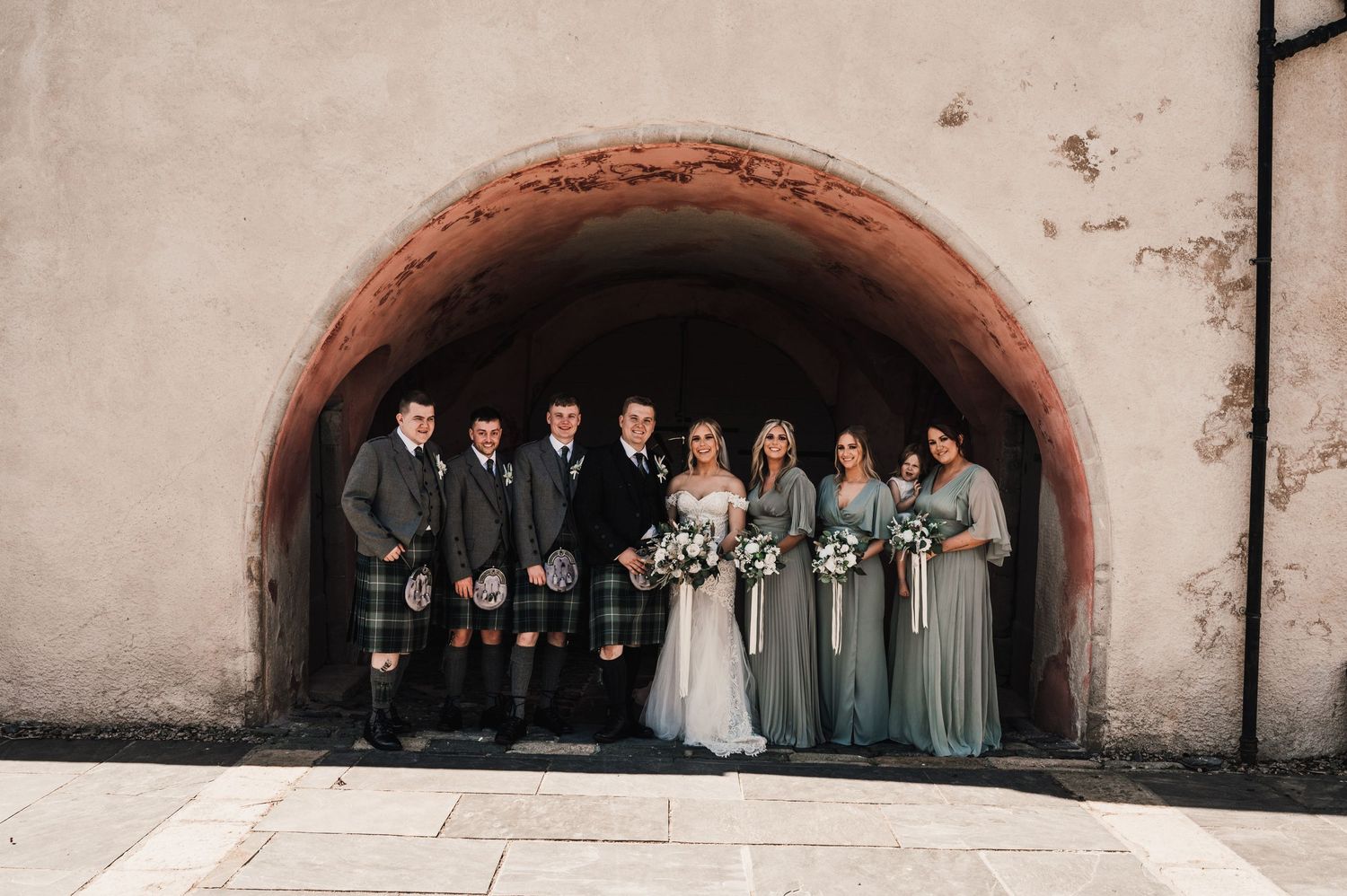 Wedding party with bridesmaids in sage dresses and groomsmen in kilts posed under stone arch.