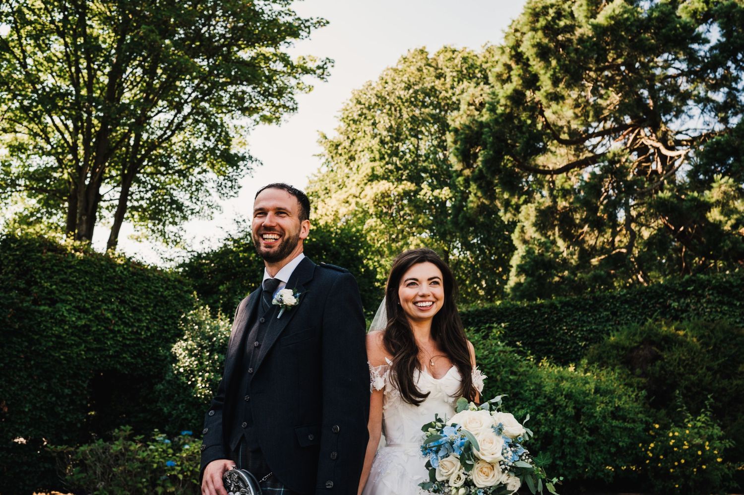 Candid garden wedding portrait with Scottish groom's attire and elegant bridal bouquet.