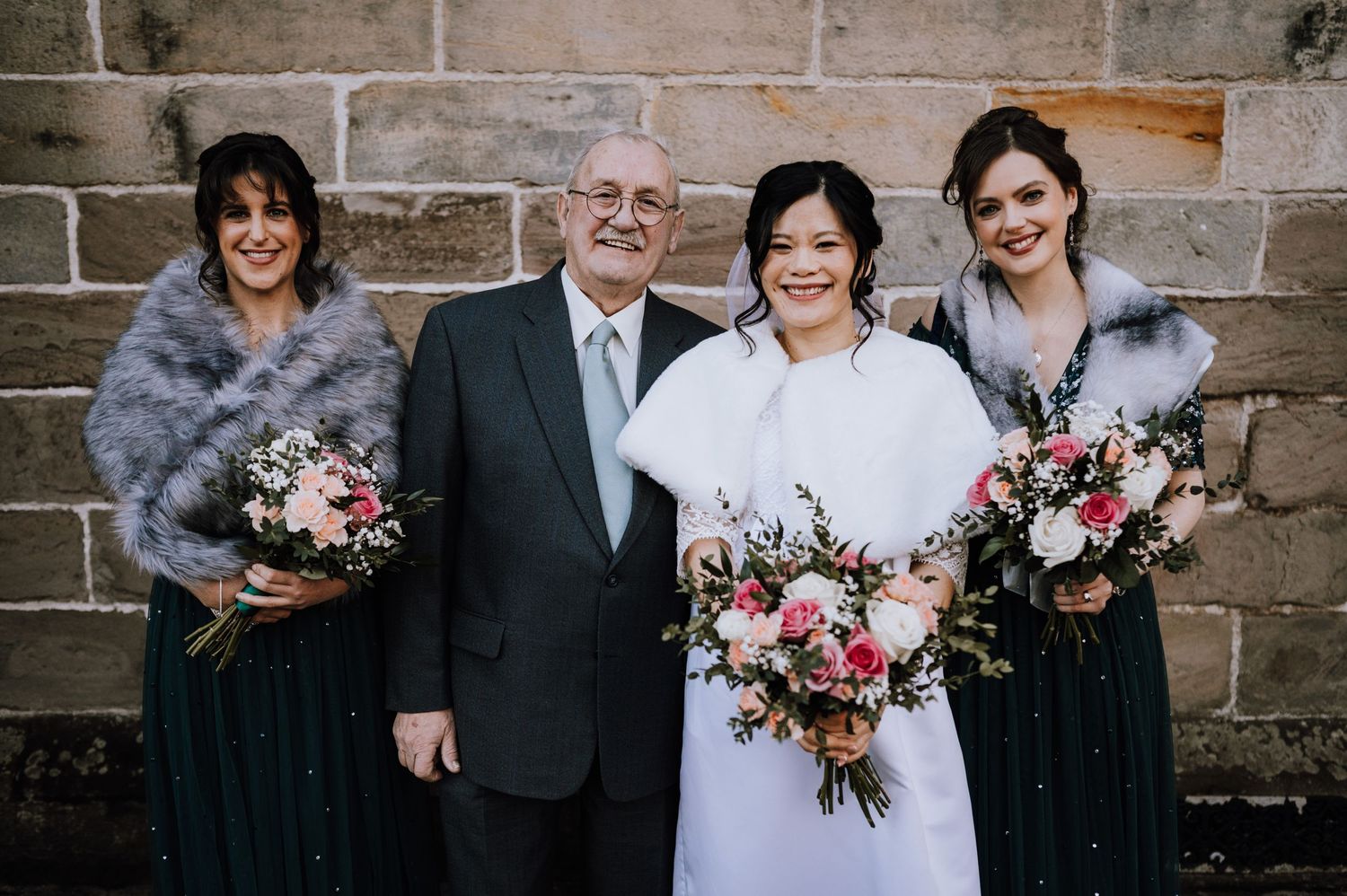 Wedding party poses against stone wall with elegant floral bouquets and fur wraps.