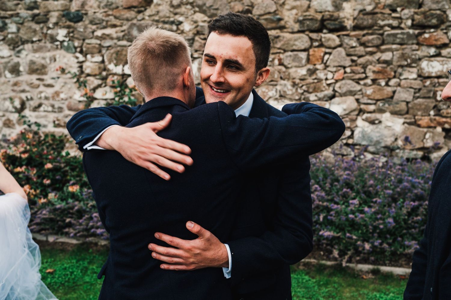Two people share emotional embrace during outdoor wedding celebration near stone wall.