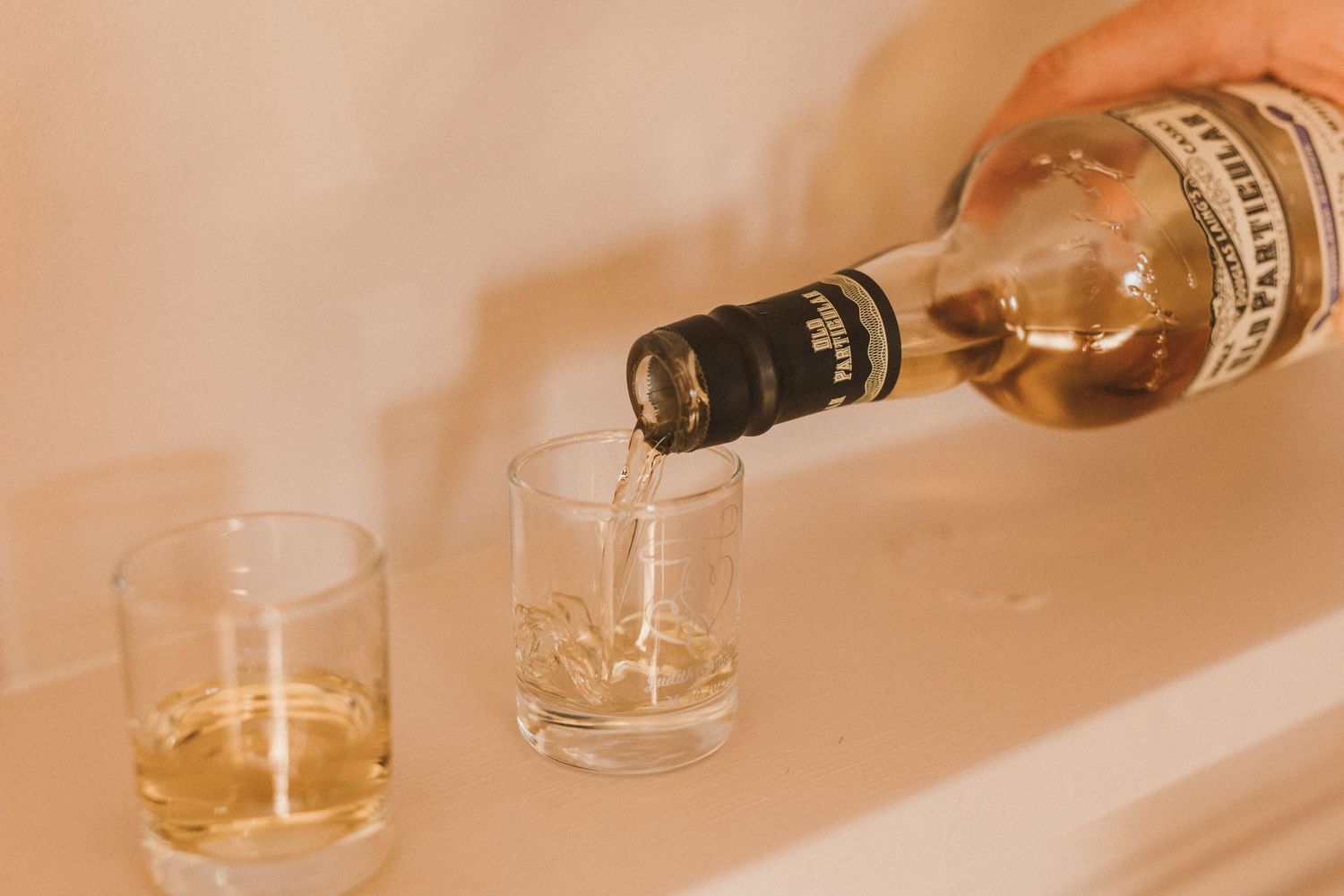 Bottle of whiskey being poured into two crystal glasses on a beige surface.