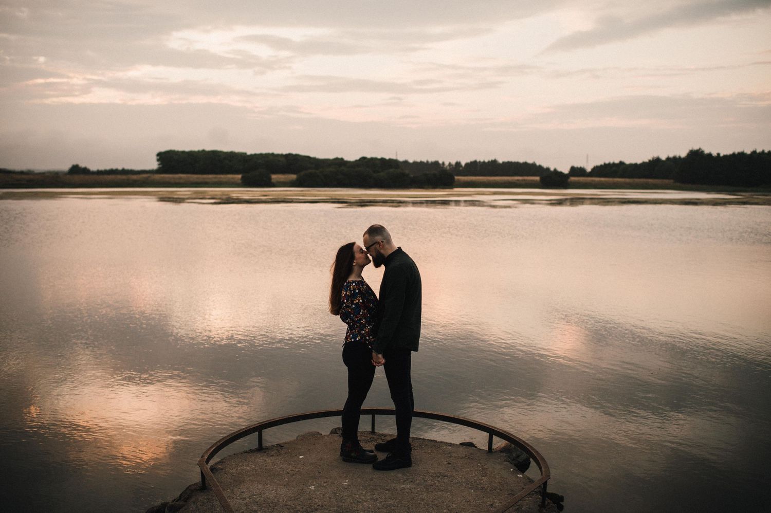 A couple embraces on a platform overlooking a serene lake at sunset.