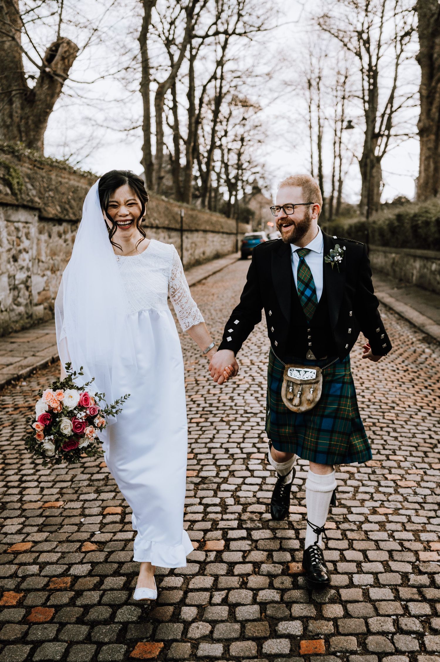 A happy couple walks along a stone-walled path, the bride with flowers and groom in traditional Scottish dress.