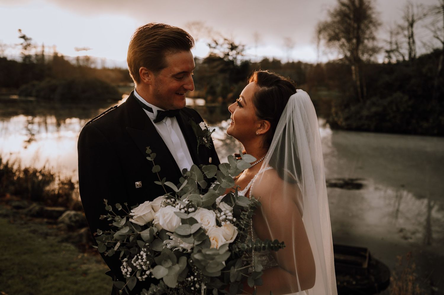 A wedding couple laughing together by a lake at sunset with a beautiful floral arrangement.