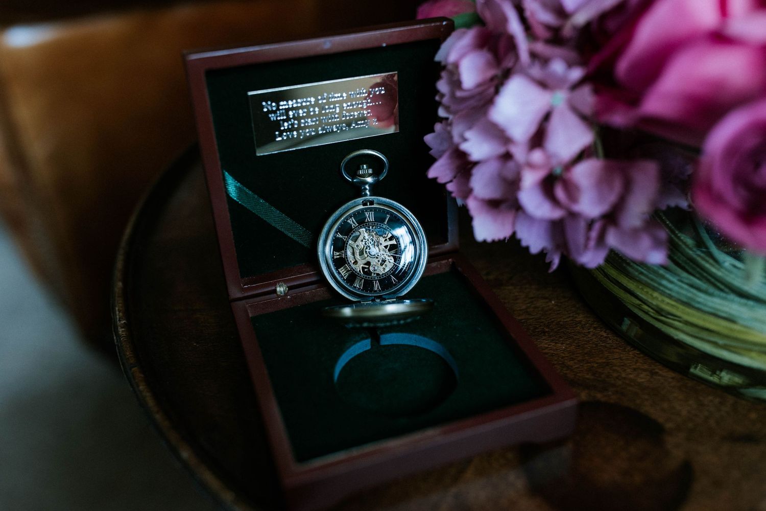 Wedding accessories including pocket watch and pink flowers arranged on dark wooden surface.