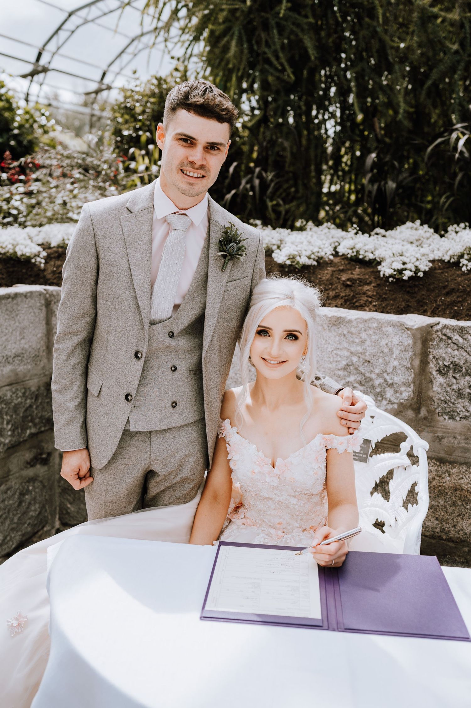 Newlyweds pose at their signing table with a purple wedding folder in a greenhouse setting.