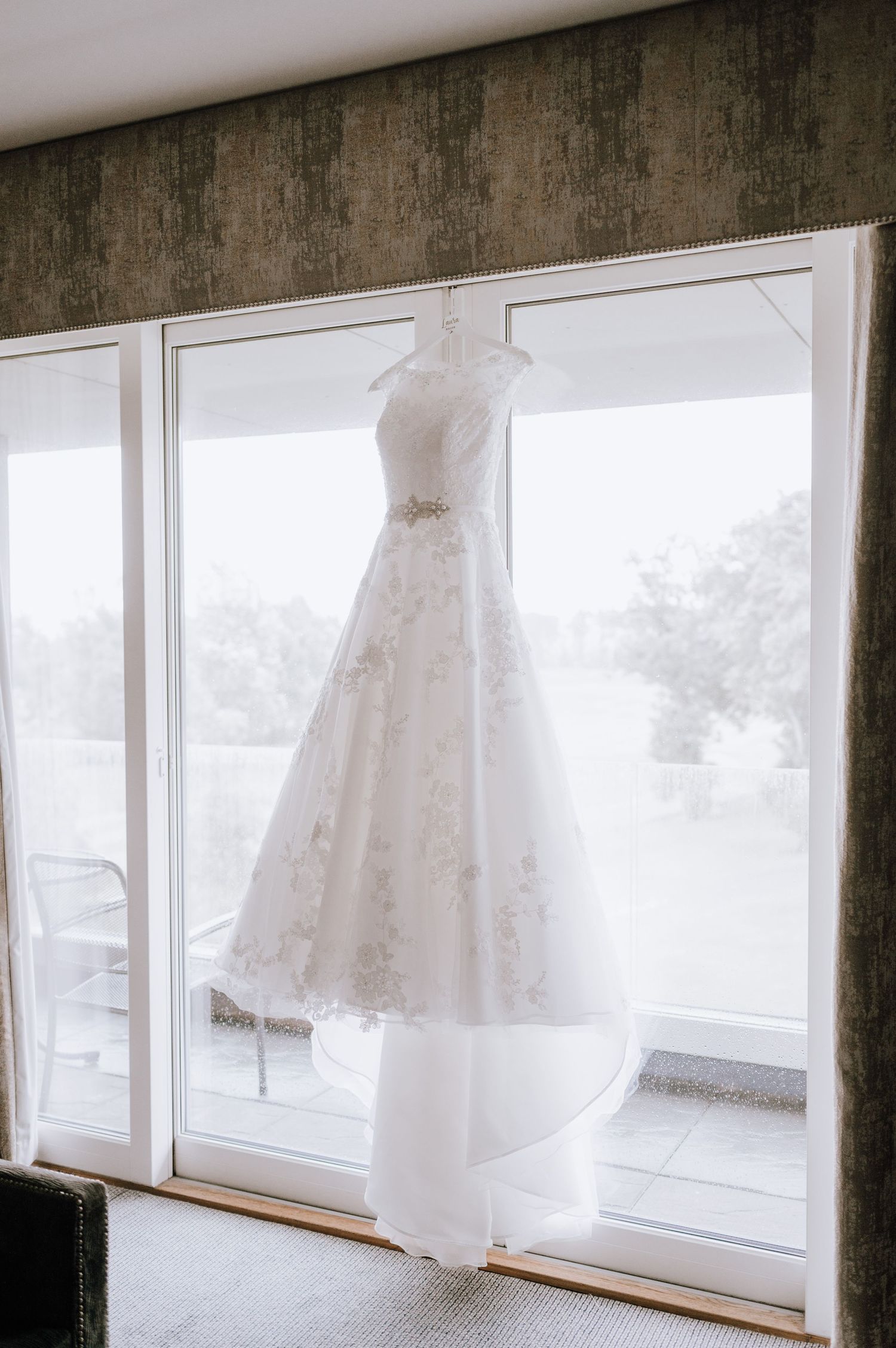 A white lace wedding gown hangs elegantly against large windows with natural light.
