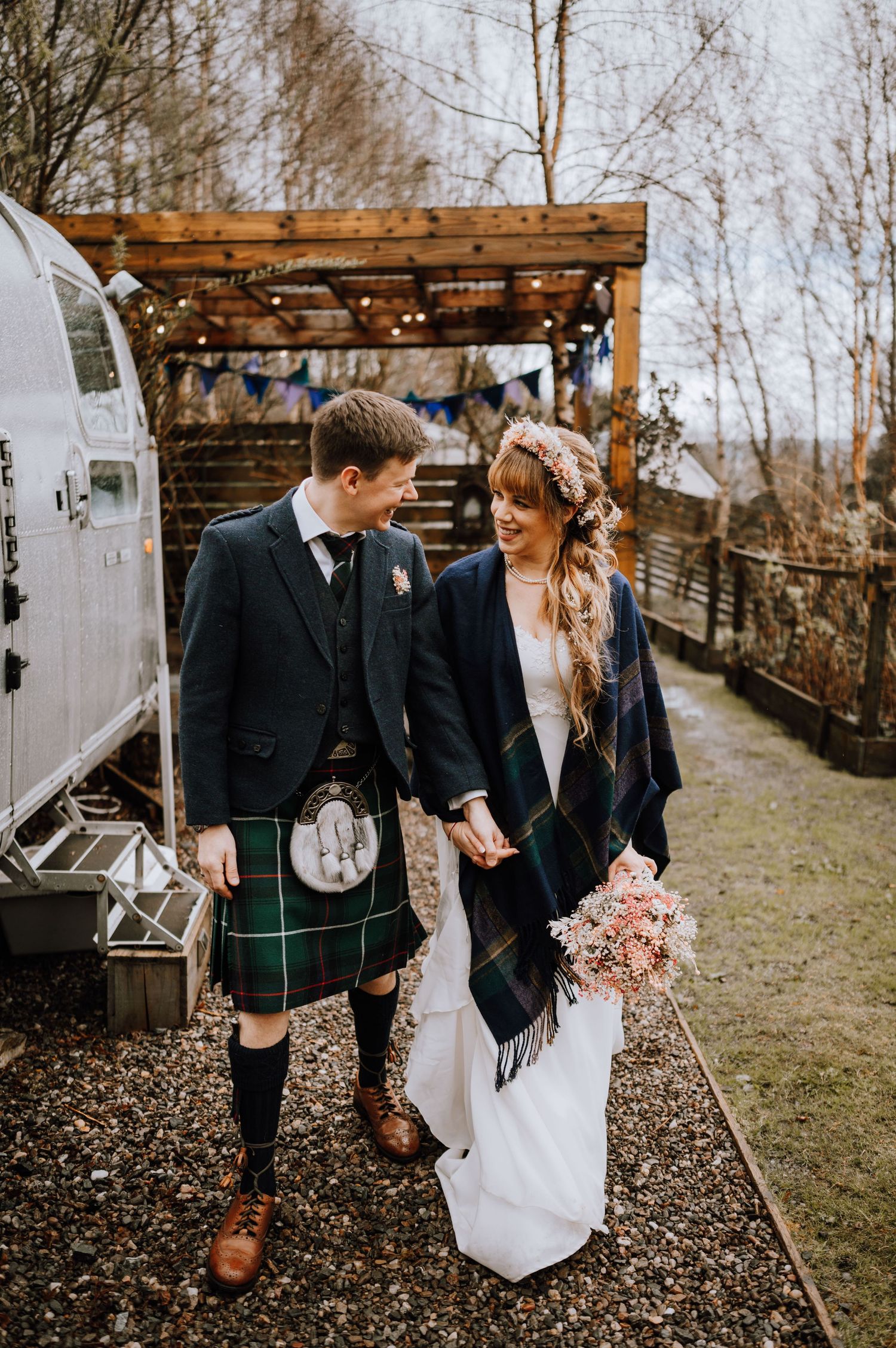 Couple in Scottish wedding attire walking together near vintage camper with string lights overhead.
