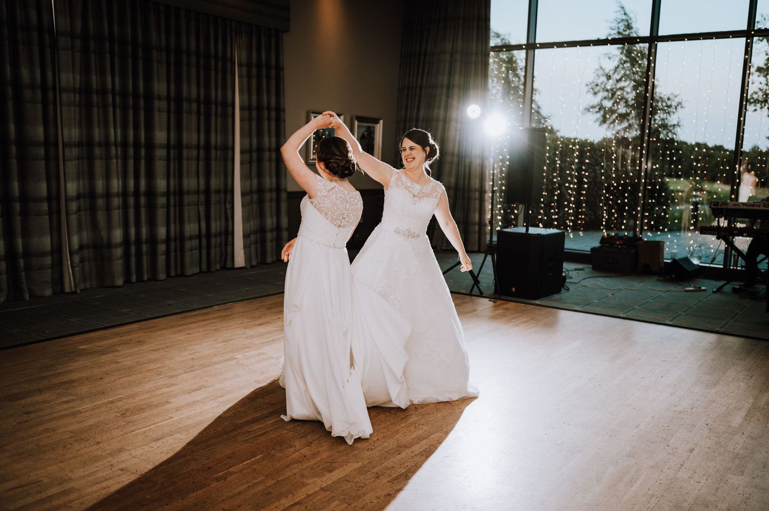Two people in white dresses dance together in a reception venue with fairy lights in the background.
