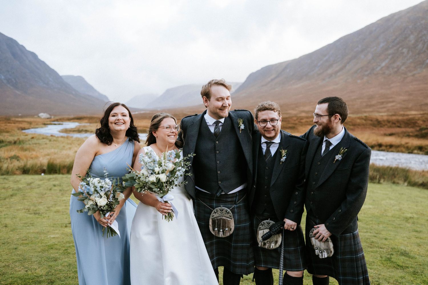 Wedding party in traditional Scottish attire poses against dramatic mountain landscape.