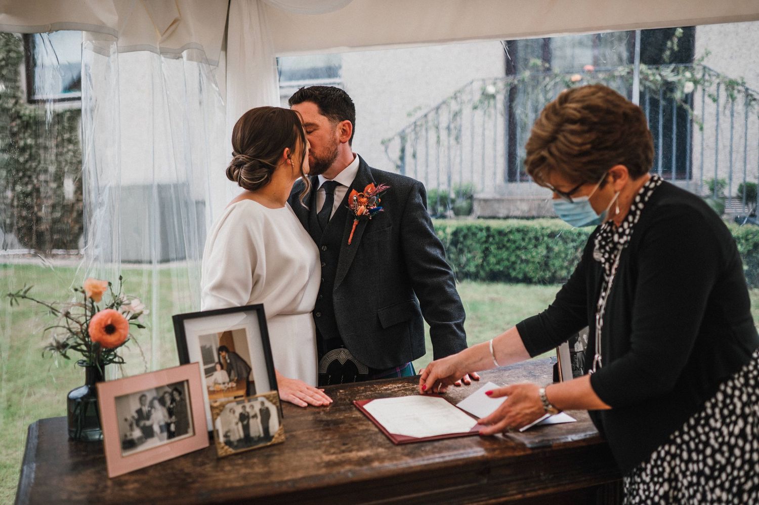 Couple signing marriage documents at rustic table with framed photos.