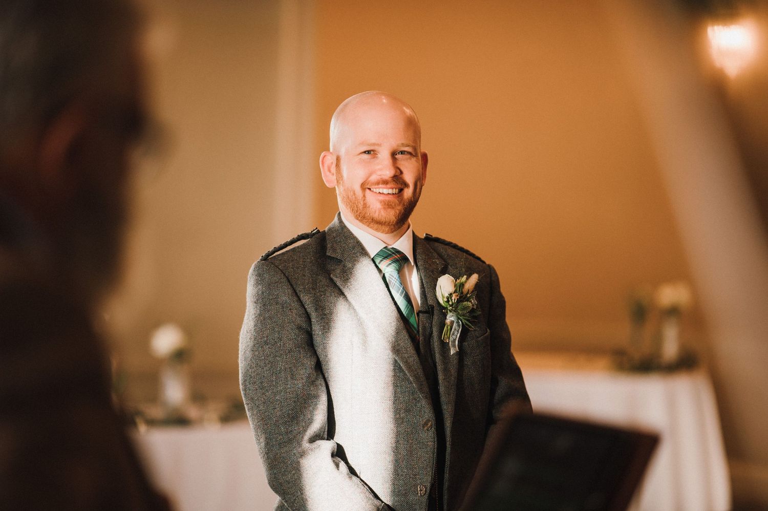 Wedding portrait showing someone in a grey suit with boutonniere smiling in warm indoor lighting.