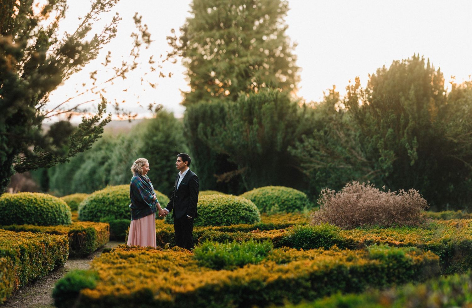 A couple stands together in a manicured garden with trimmed hedges at sunset.