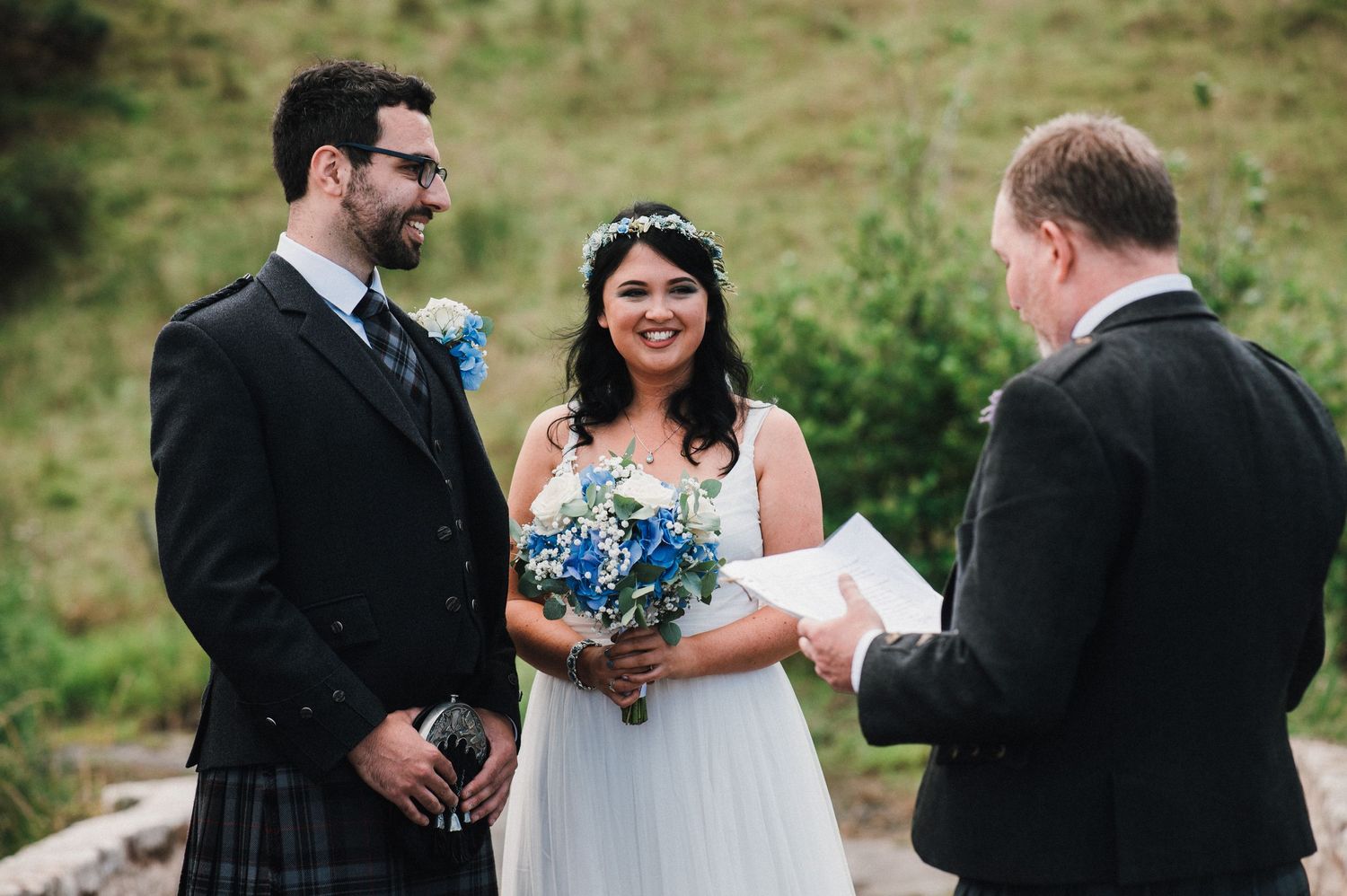 Outdoor wedding ceremony with traditional Scottish kilts against green hillside.