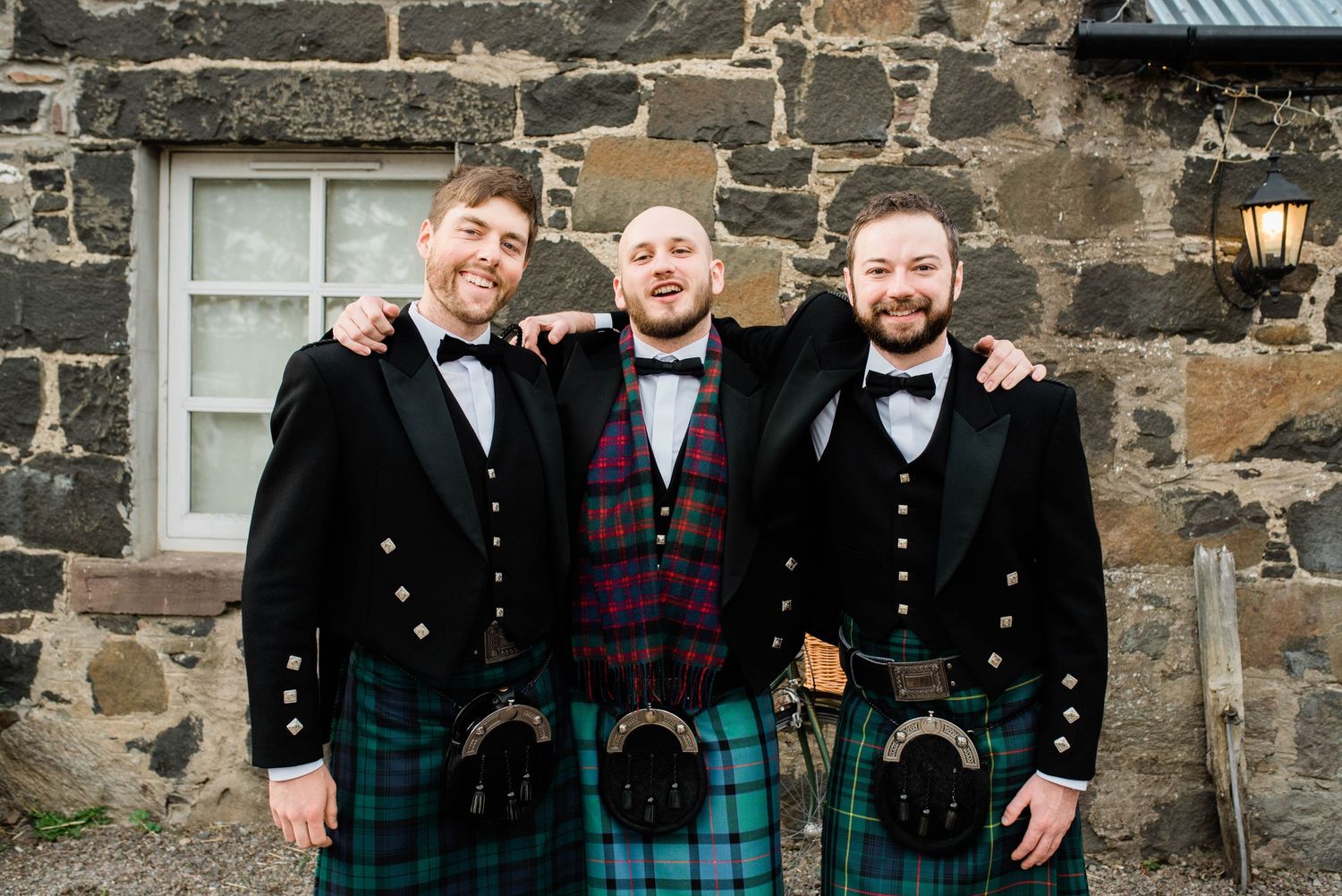 Three groomsmen in traditional Scottish kilts pose against stone wall.