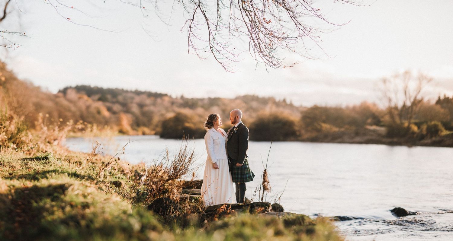 Wide landscape wedding photo of couple by riverside in autumn setting.