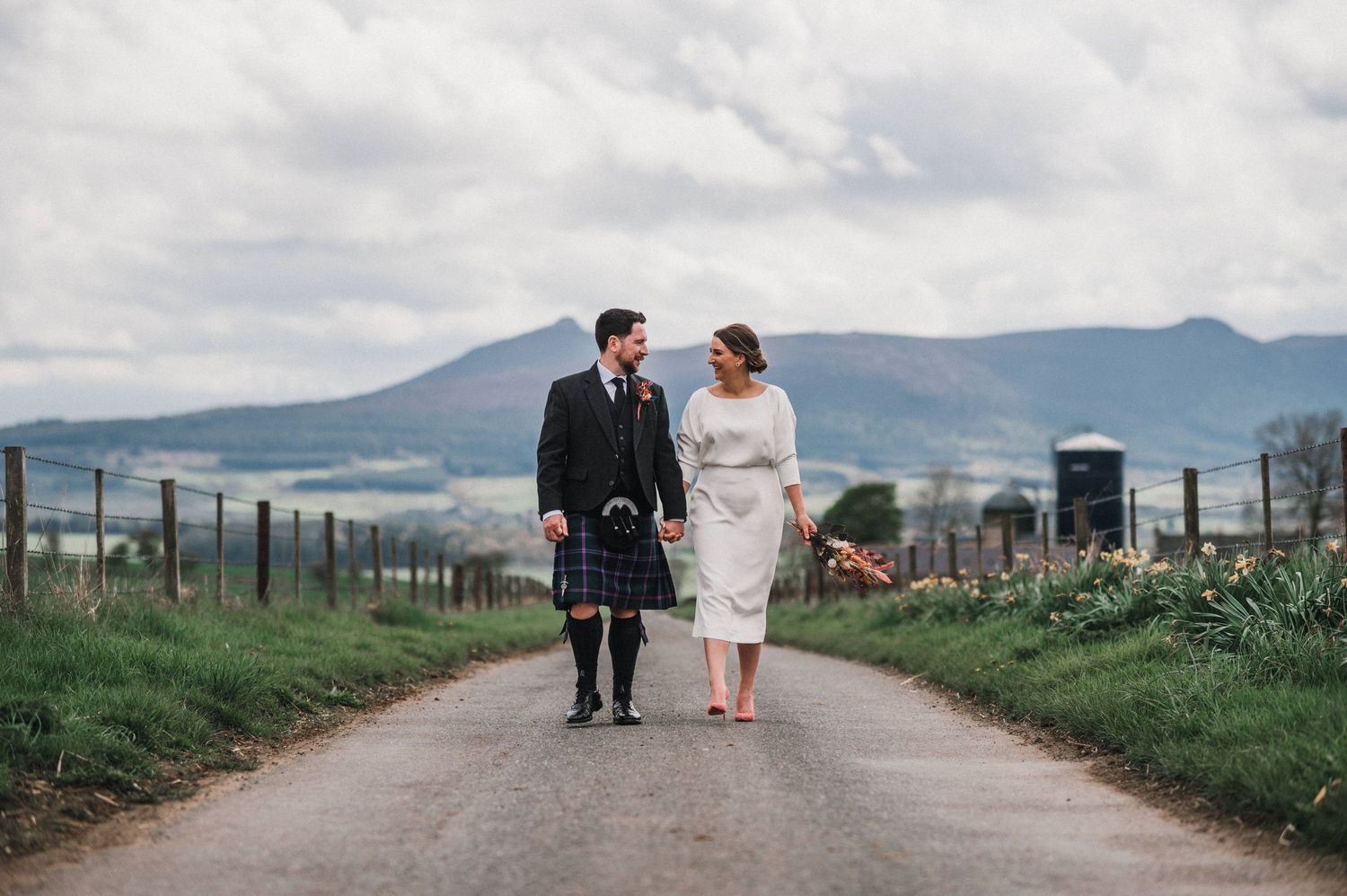 A couple walks together on a country road with mountains in the distance, one wearing traditional Scottish attire.