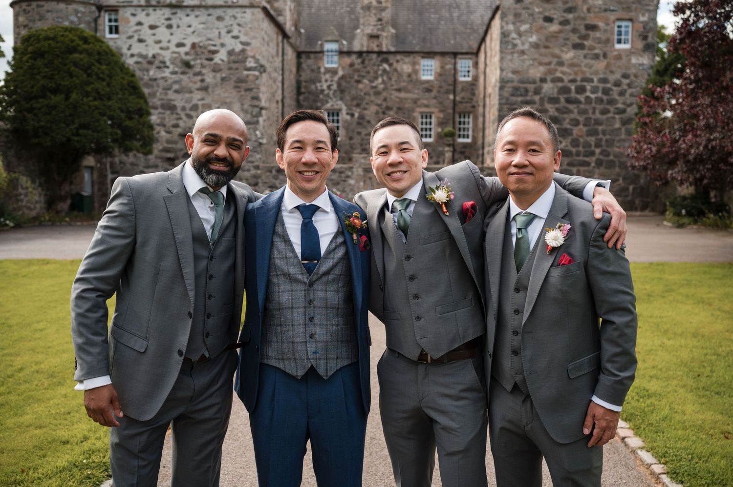 Group photo of wedding party members in formal attire outside stone building.