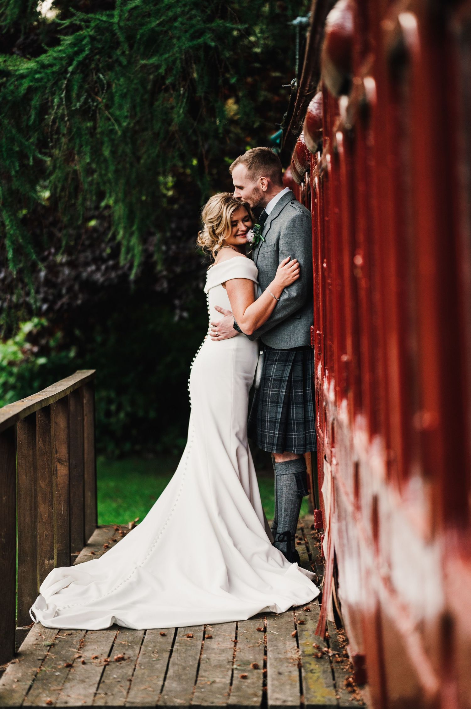 Romantic wedding portrait against a red wall with bride in flowing white dress and groom in traditional Scottish kilt.