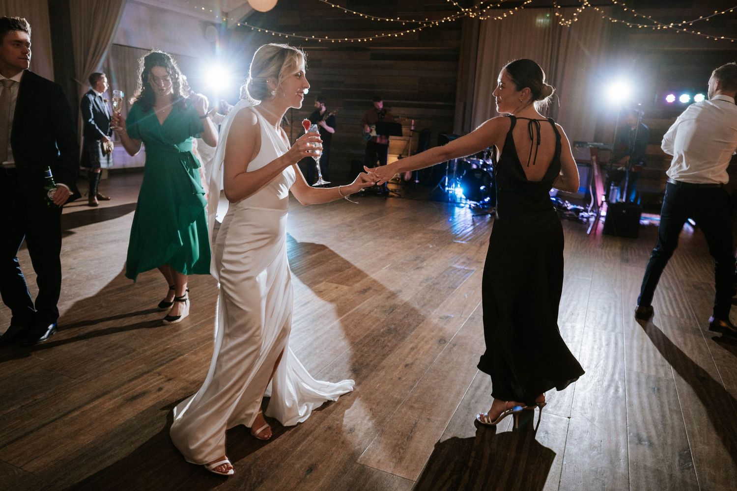 Wedding guests dancing on wooden floor under string lights at reception.