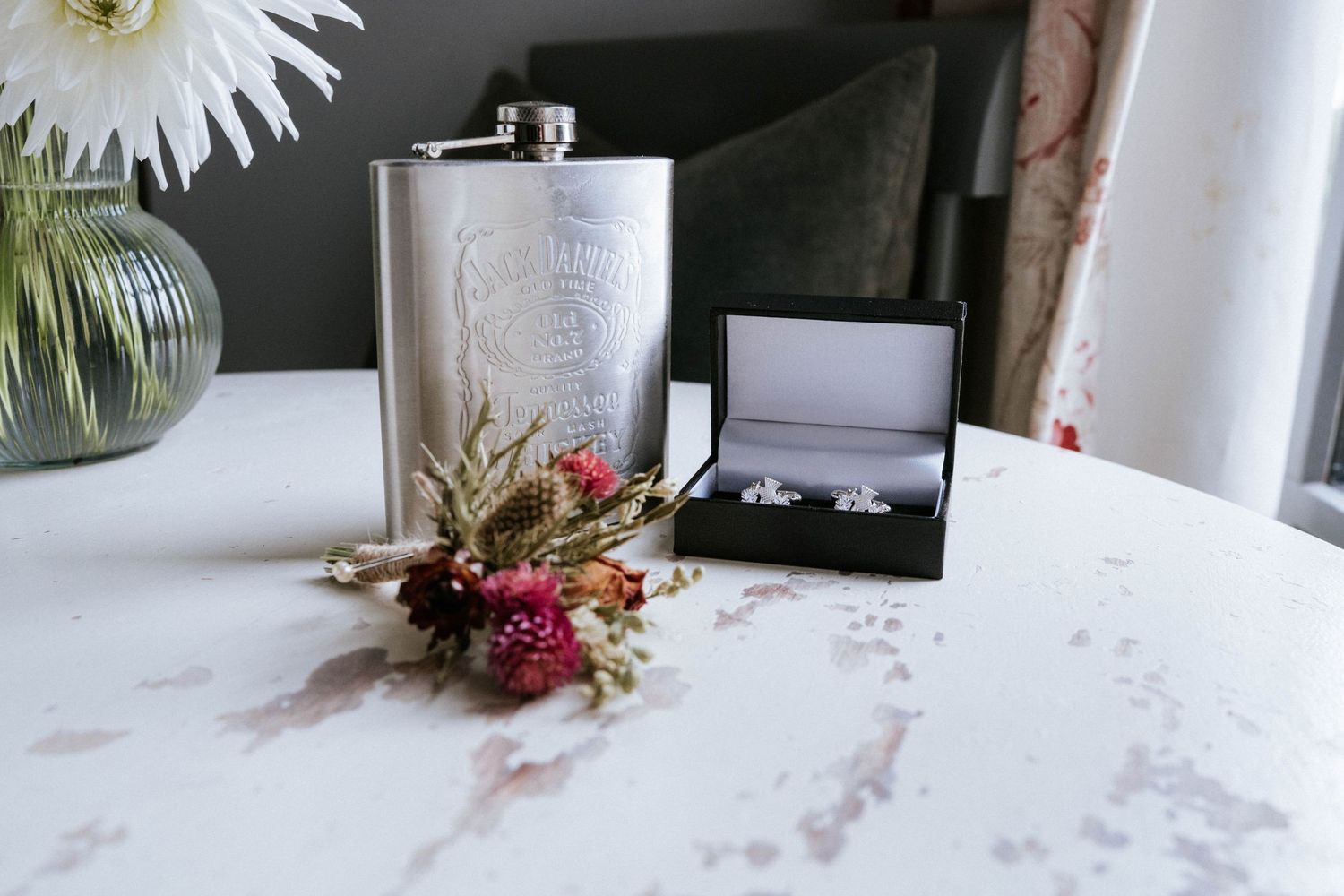 Wedding details with silver flask and ring box on marble surface with delicate floral accents.