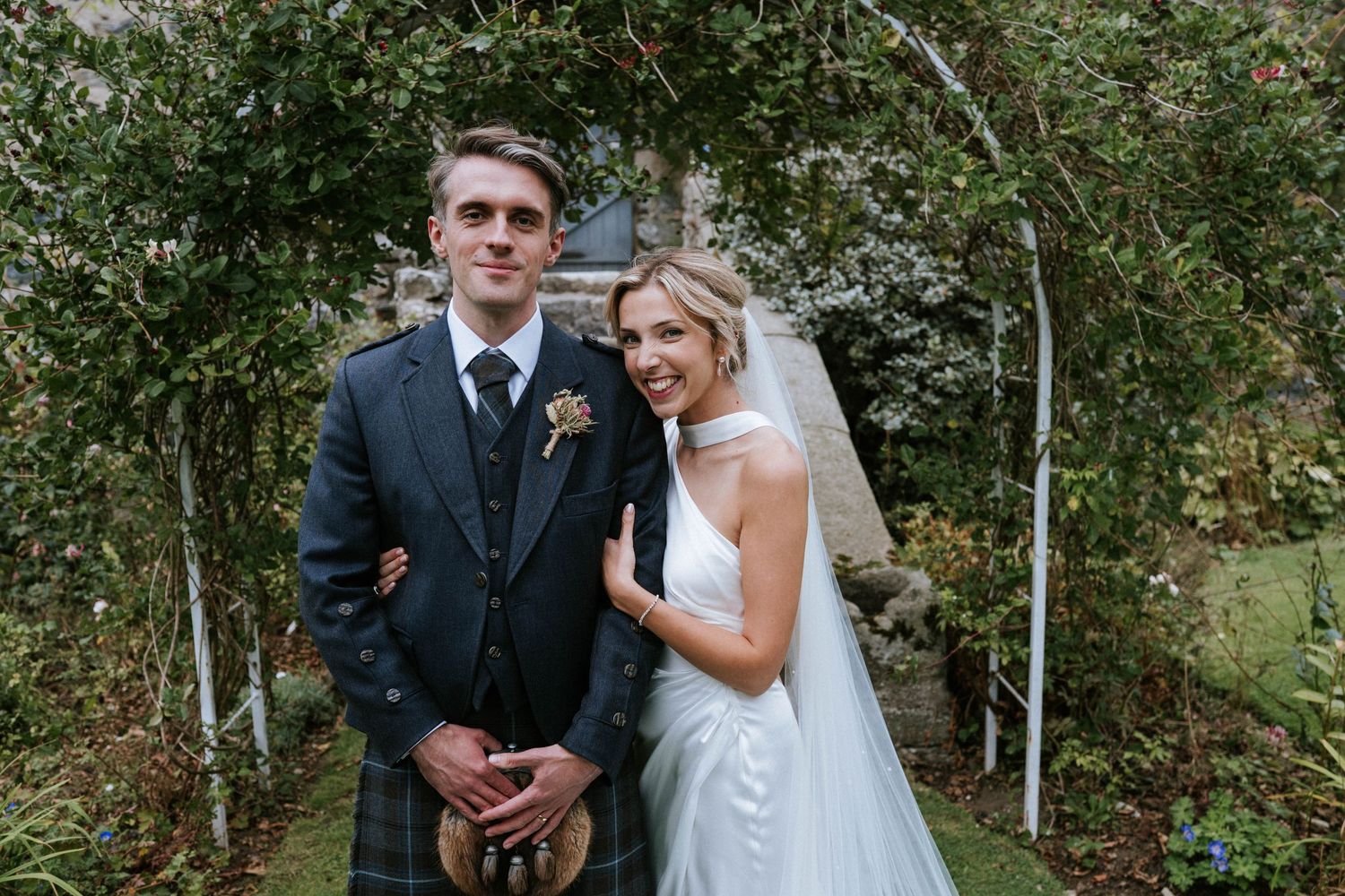 Newlyweds pose under garden arch with groom wearing traditional Scottish attire.