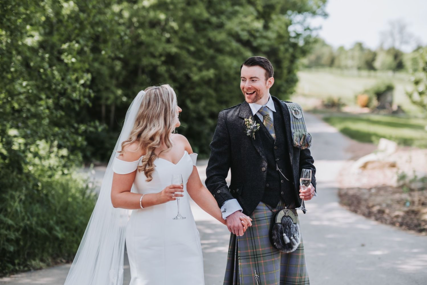 A happy bride and groom share a candid moment walking together outdoors, with the groom wearing a traditional Scottish kilt.