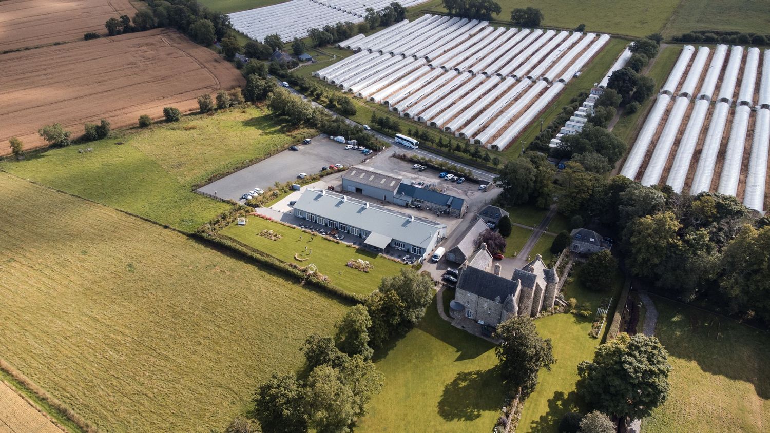 Aerial view of a large greenhouse facility with solar panels and agricultural fields surrounding a rural property.