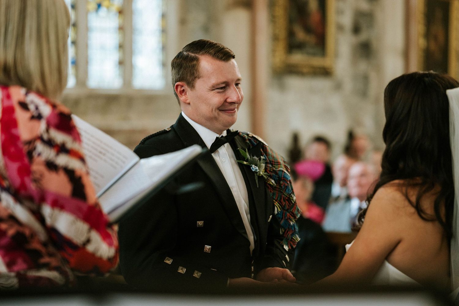 Intimate moment during wedding vows exchange in historic church interior with stained glass backdrop.