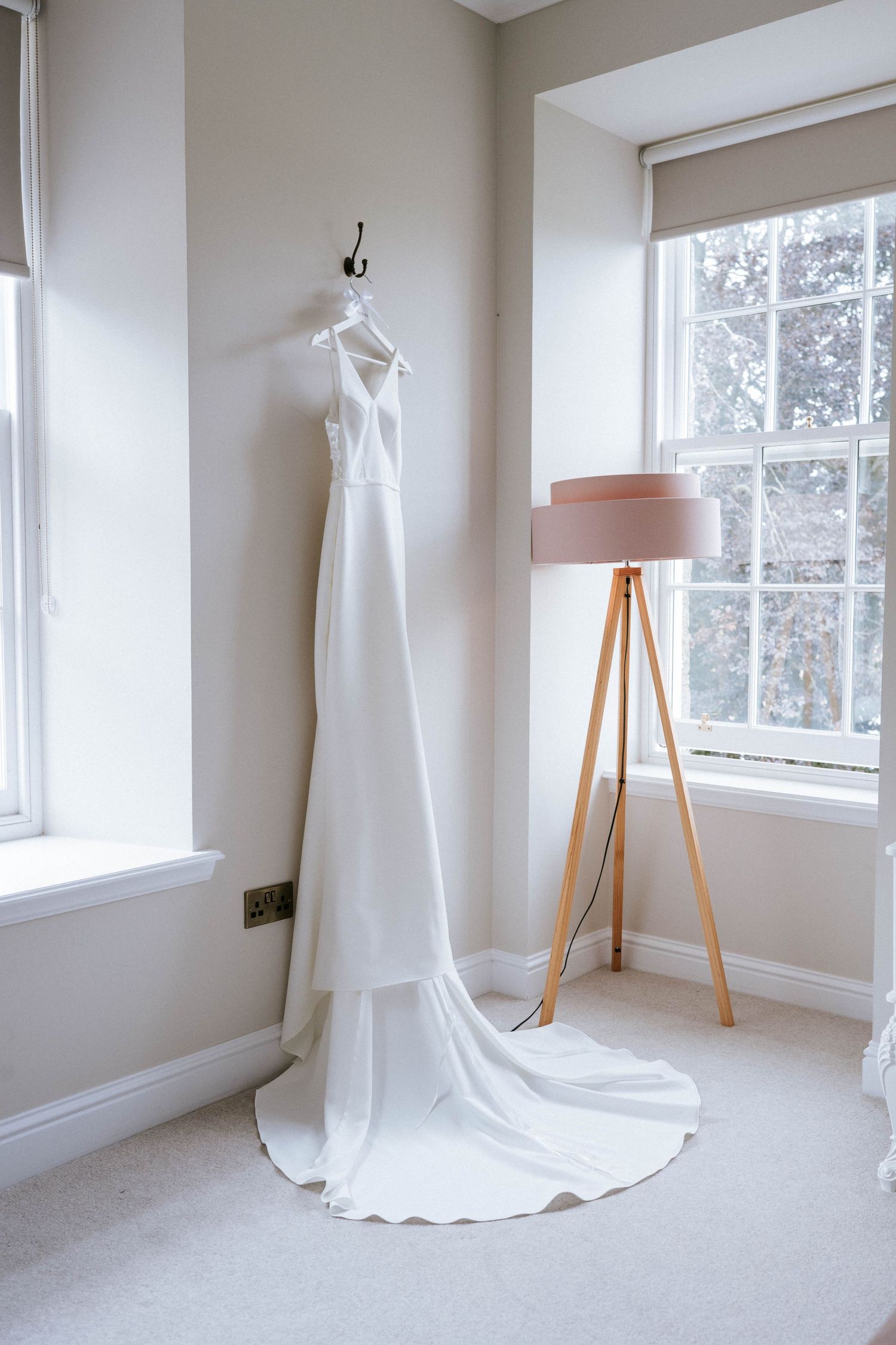 Elegant white wedding dress displayed in bright, minimalist room with natural lighting and wooden floor lamp.