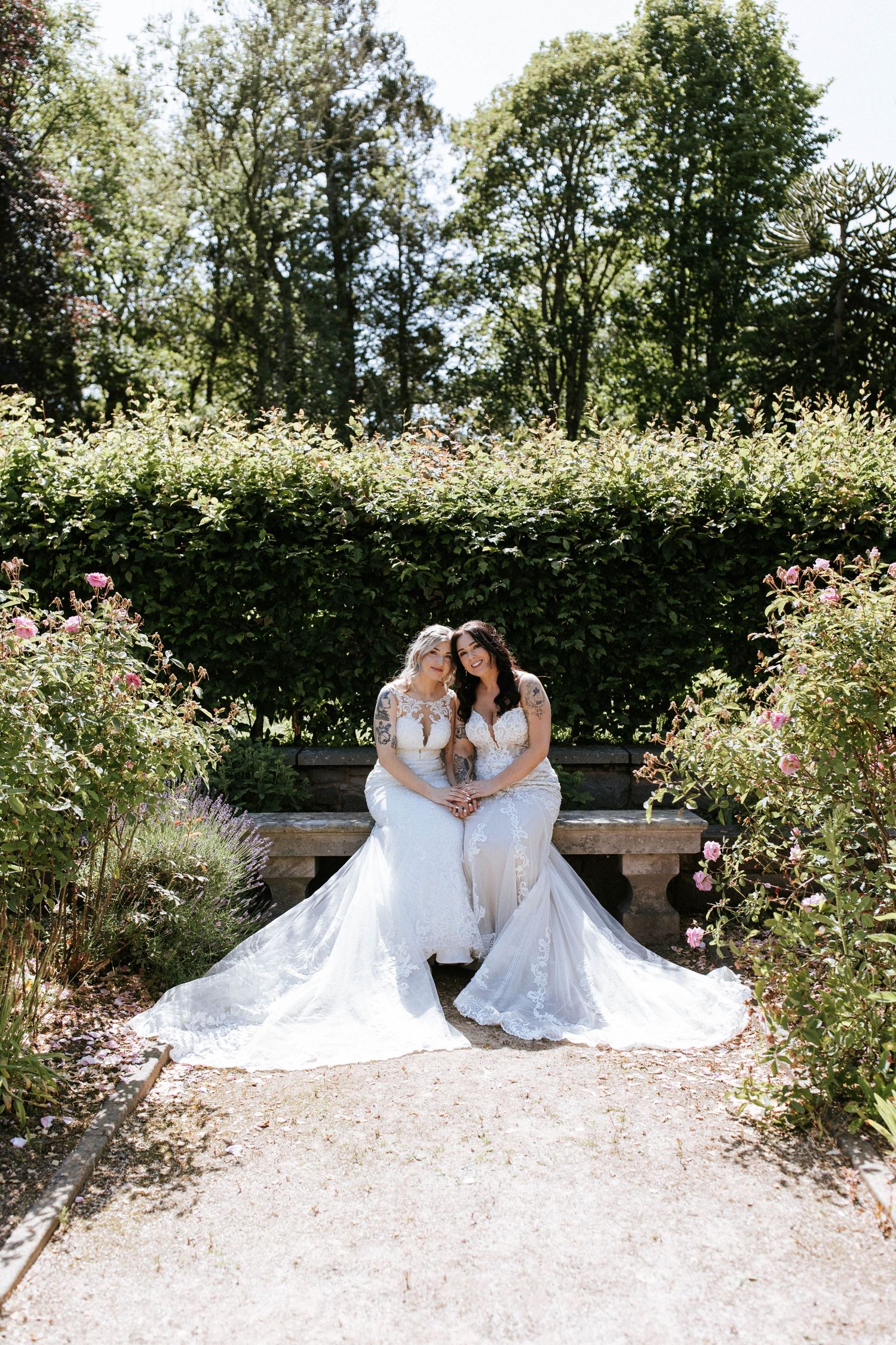 Two people in white wedding dresses sitting together on stone bench surrounded by blooming flowers.