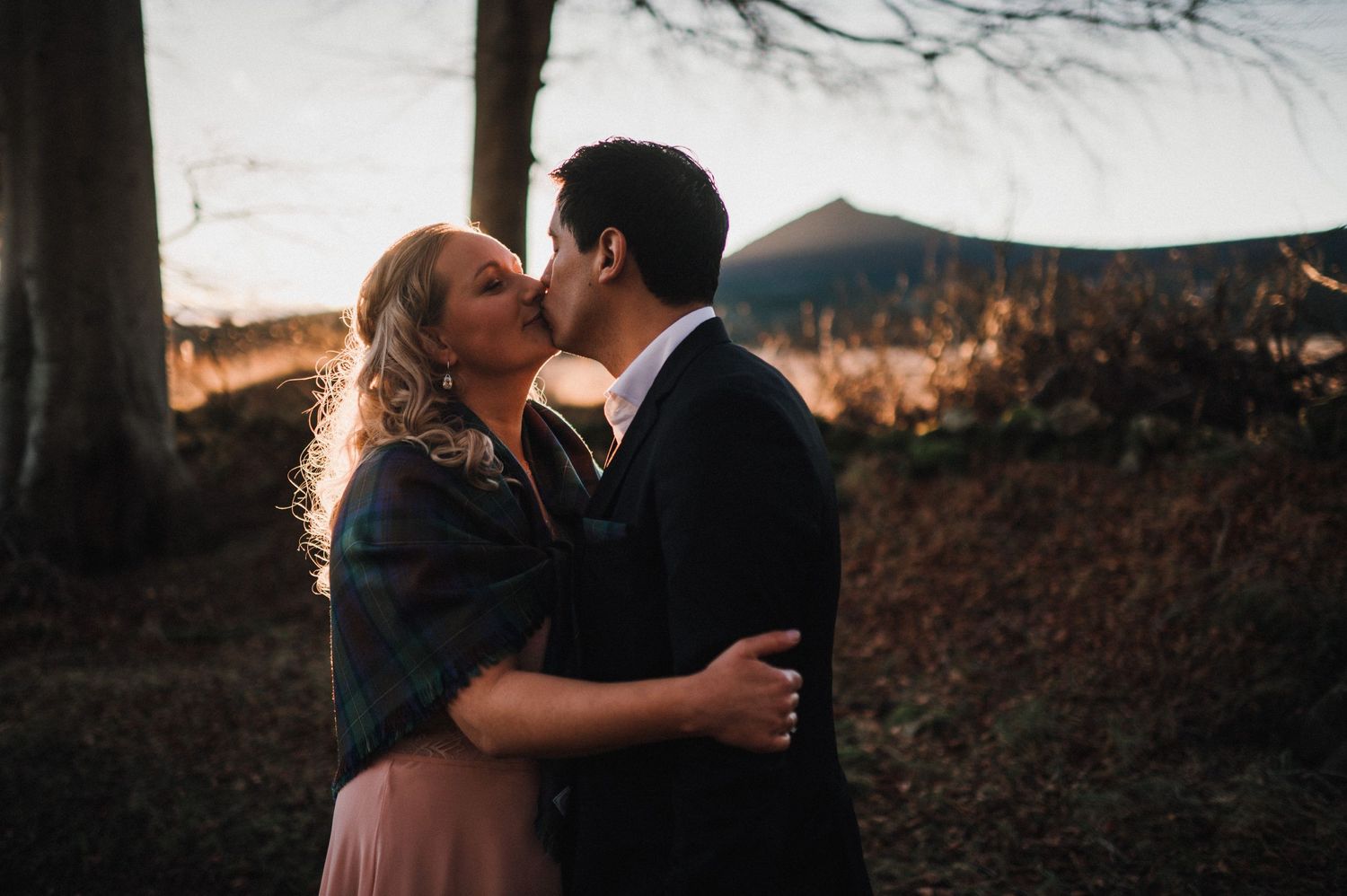 A couple embraces in warm evening light beneath tall trees.