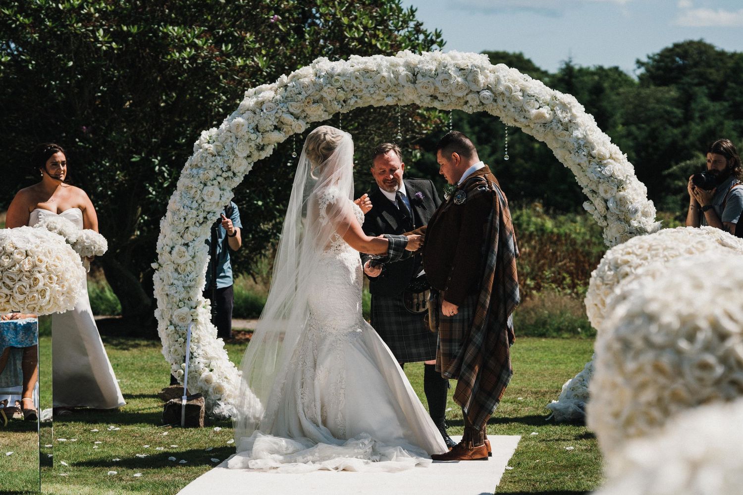 Wedding ceremony under an ornate white floral arch with the couple exchanging vows.