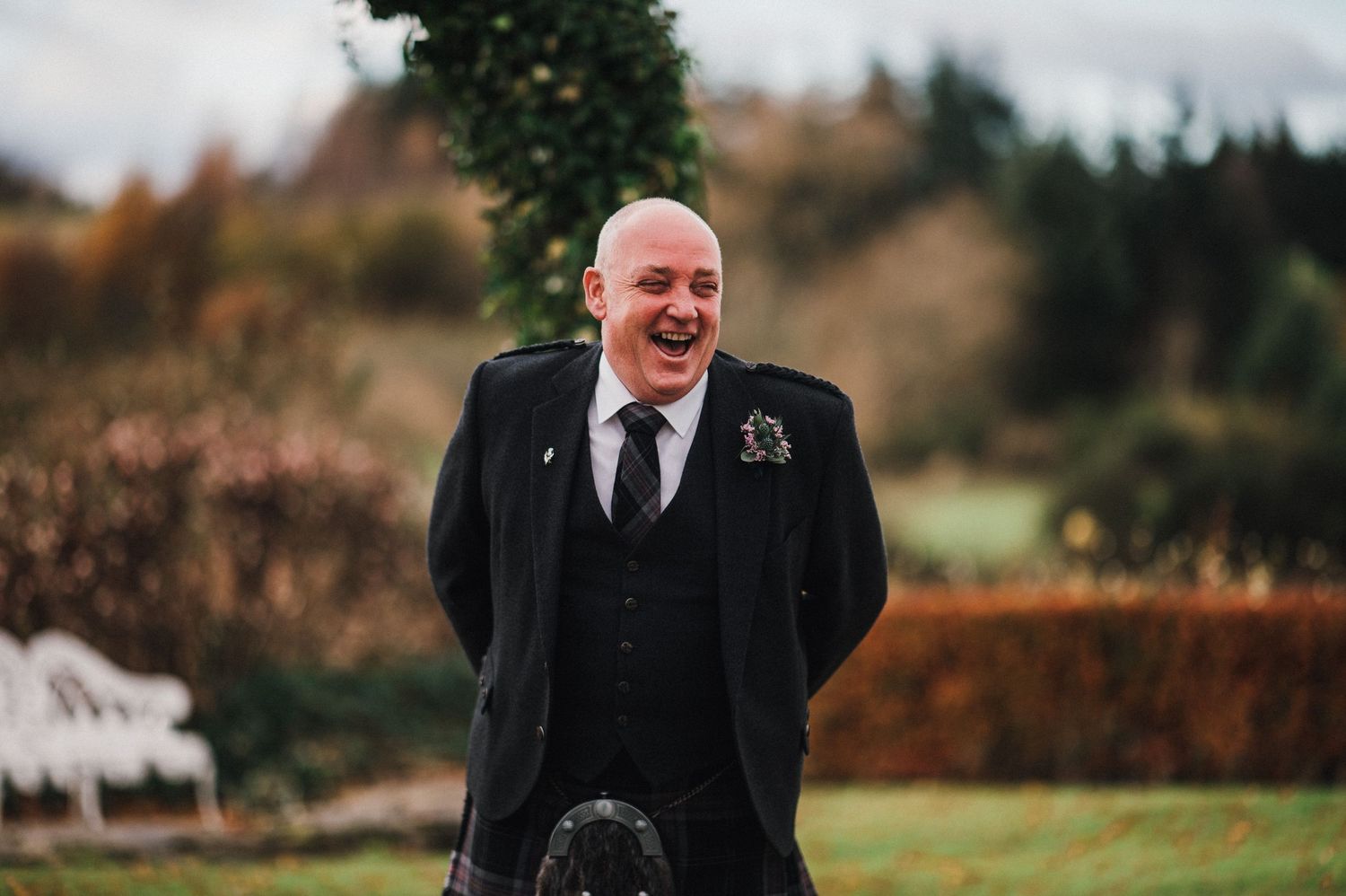 A candid moment showing someone wearing a traditional black kilt and suit jacket outdoors at a wedding event.