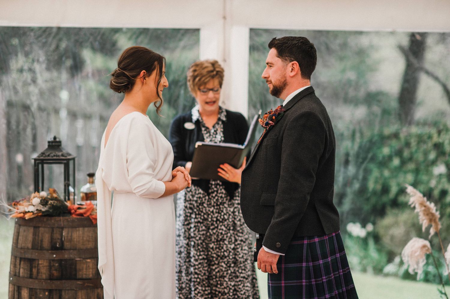 Intimate wedding ceremony taking place under a white tent with rustic barrel decor.