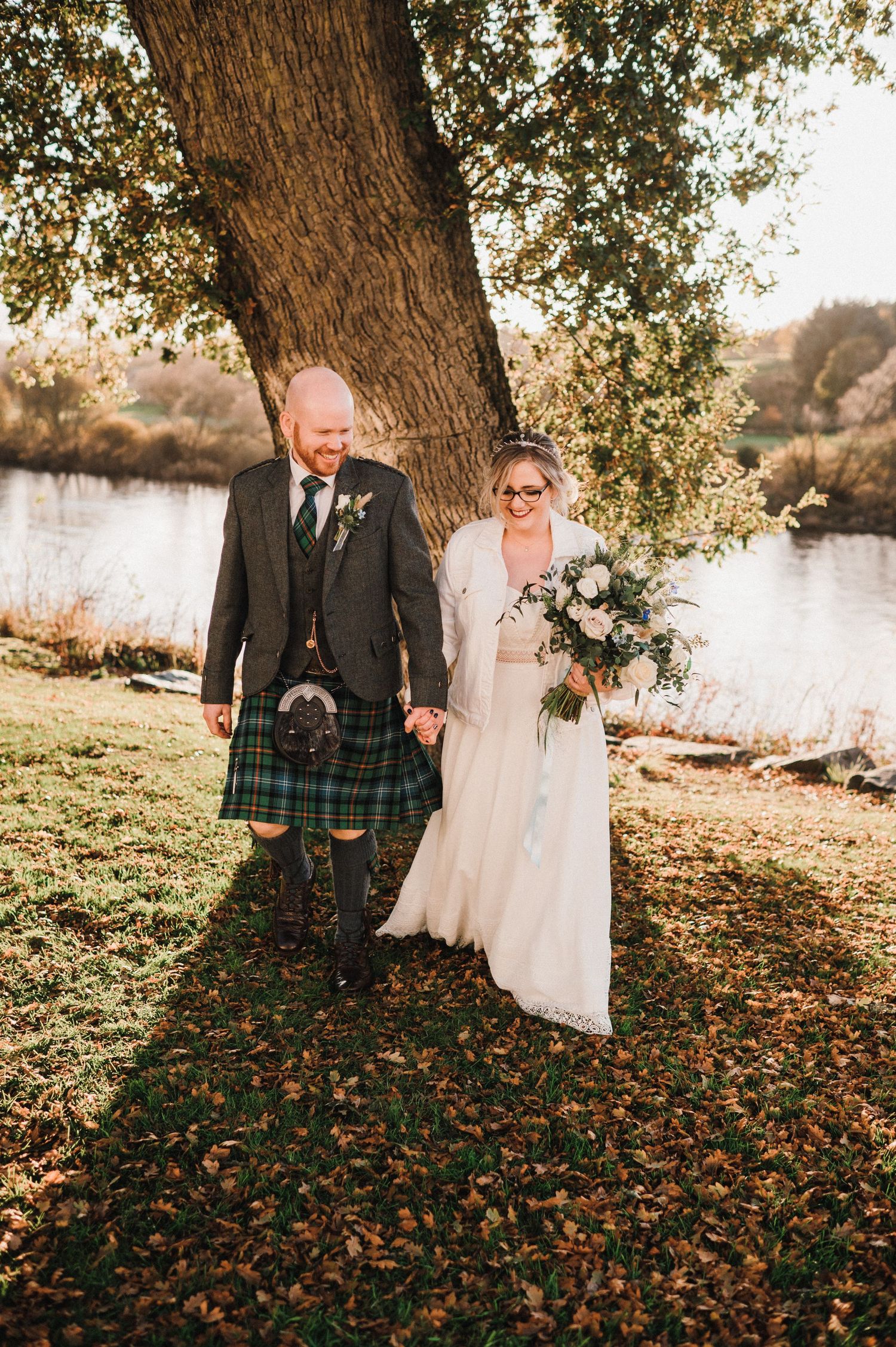 Couple walking through autumn leaves by a lake, groom wearing traditional Scottish kilt.