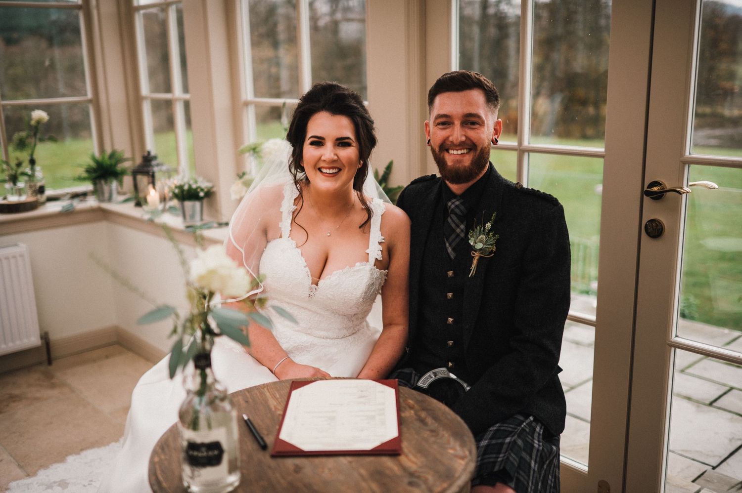 Wedding couple sits at table in sunlit conservatory with traditional Scottish attire.
