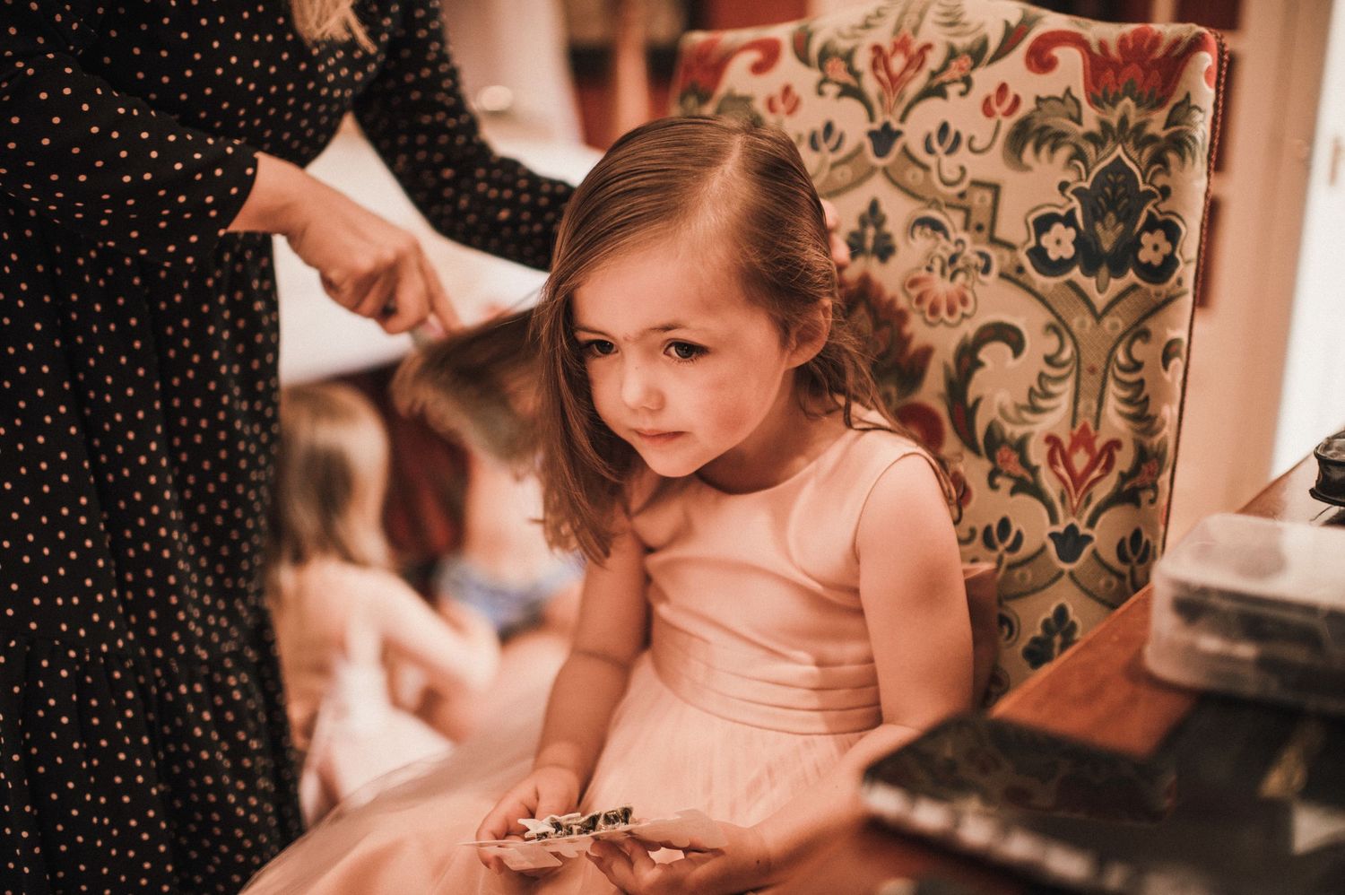 A young child in a pink dress sitting in an elegant chair during preparations.