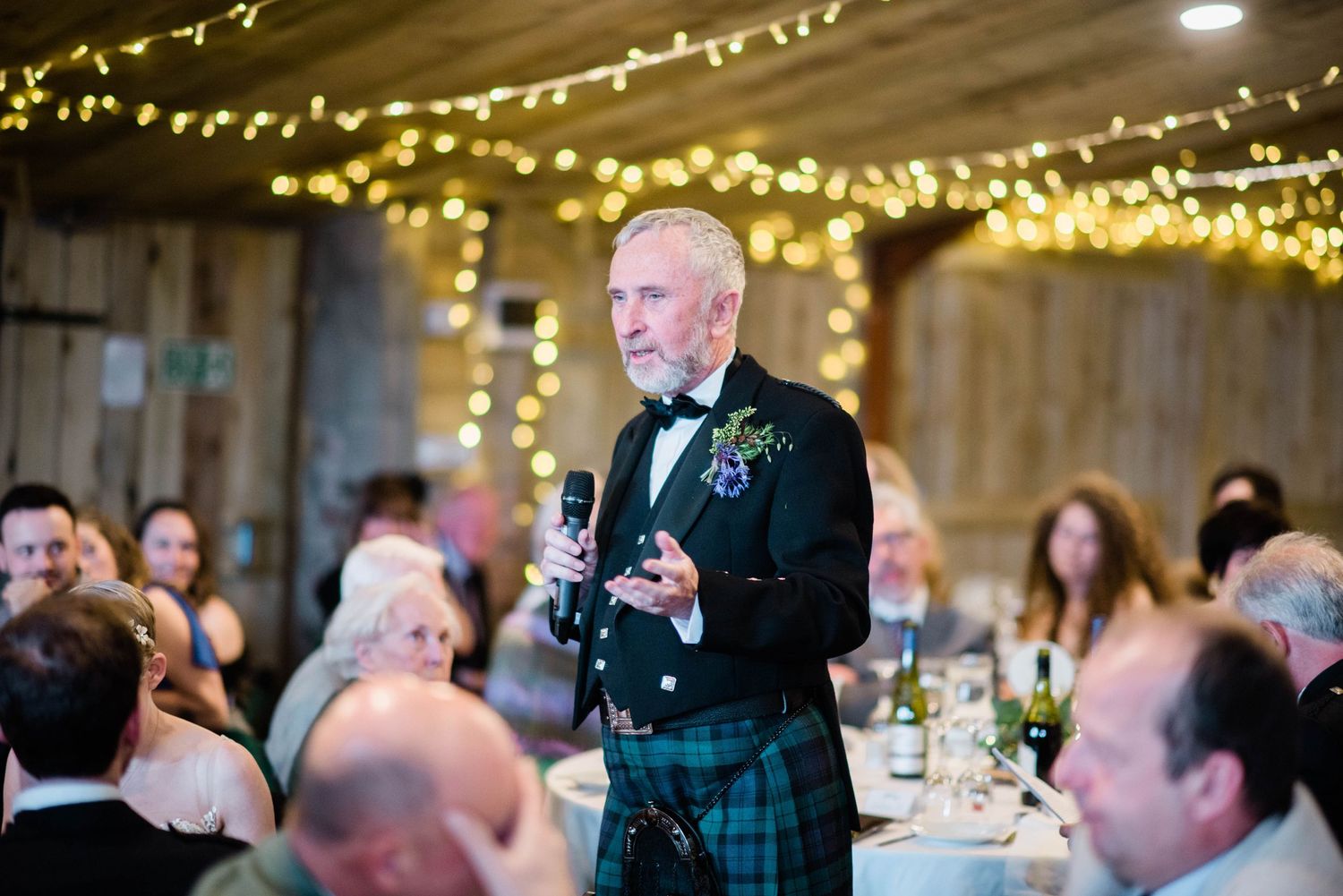 Guest wearing a traditional kilt gives a speech at a wedding reception under twinkling fairy lights.