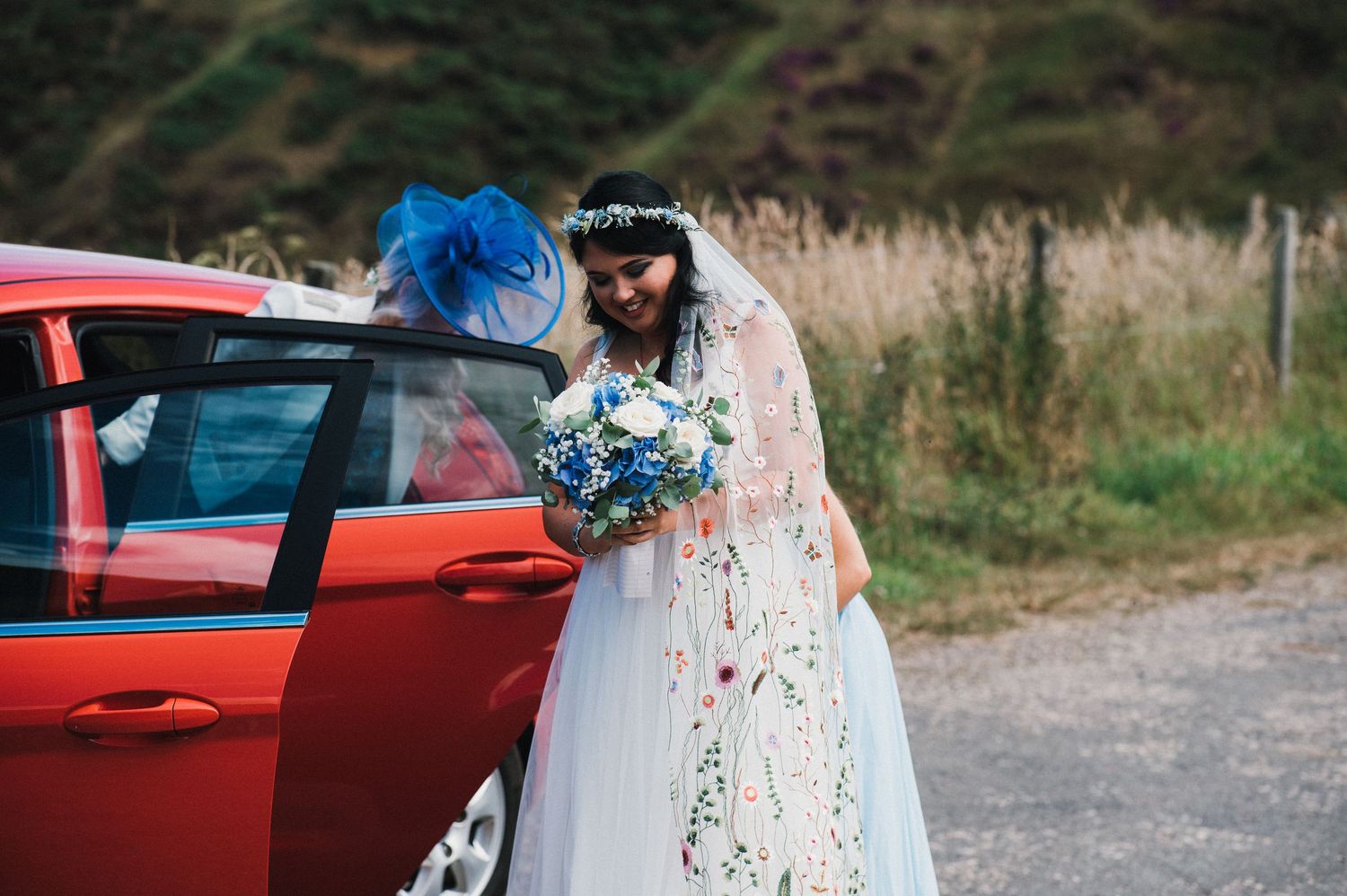 Bride in white dress holding blue bouquet stands by red car on country road.