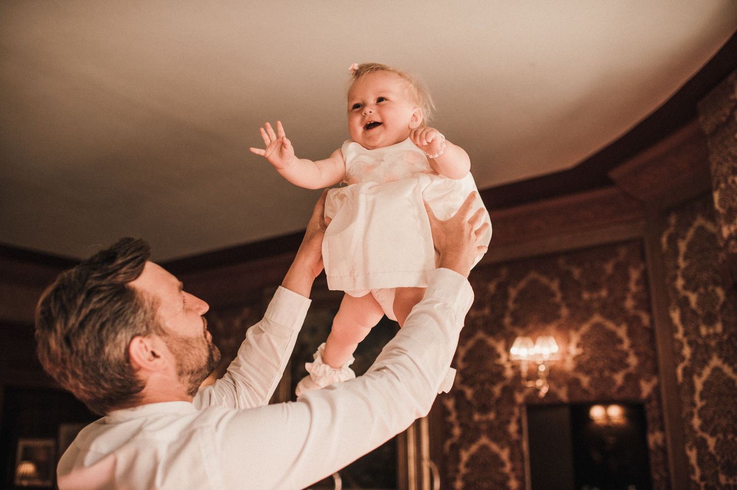 Sweet moment of parent lifting baby in white dress during wedding celebration.