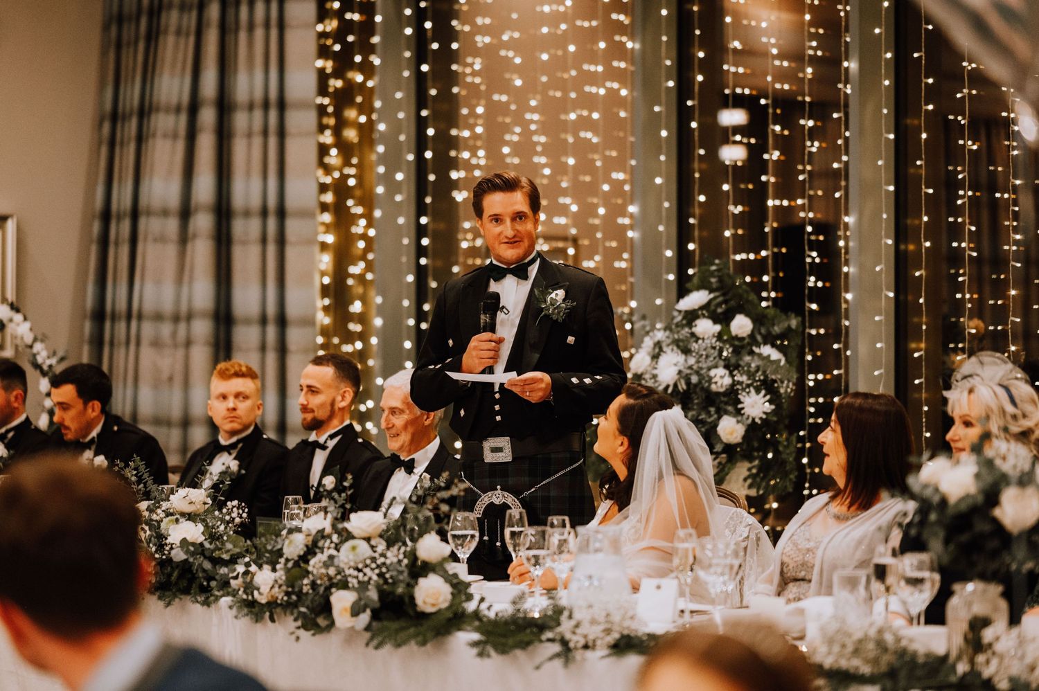 Guest delivers speech at wedding reception head table decorated with white flowers and string lights.