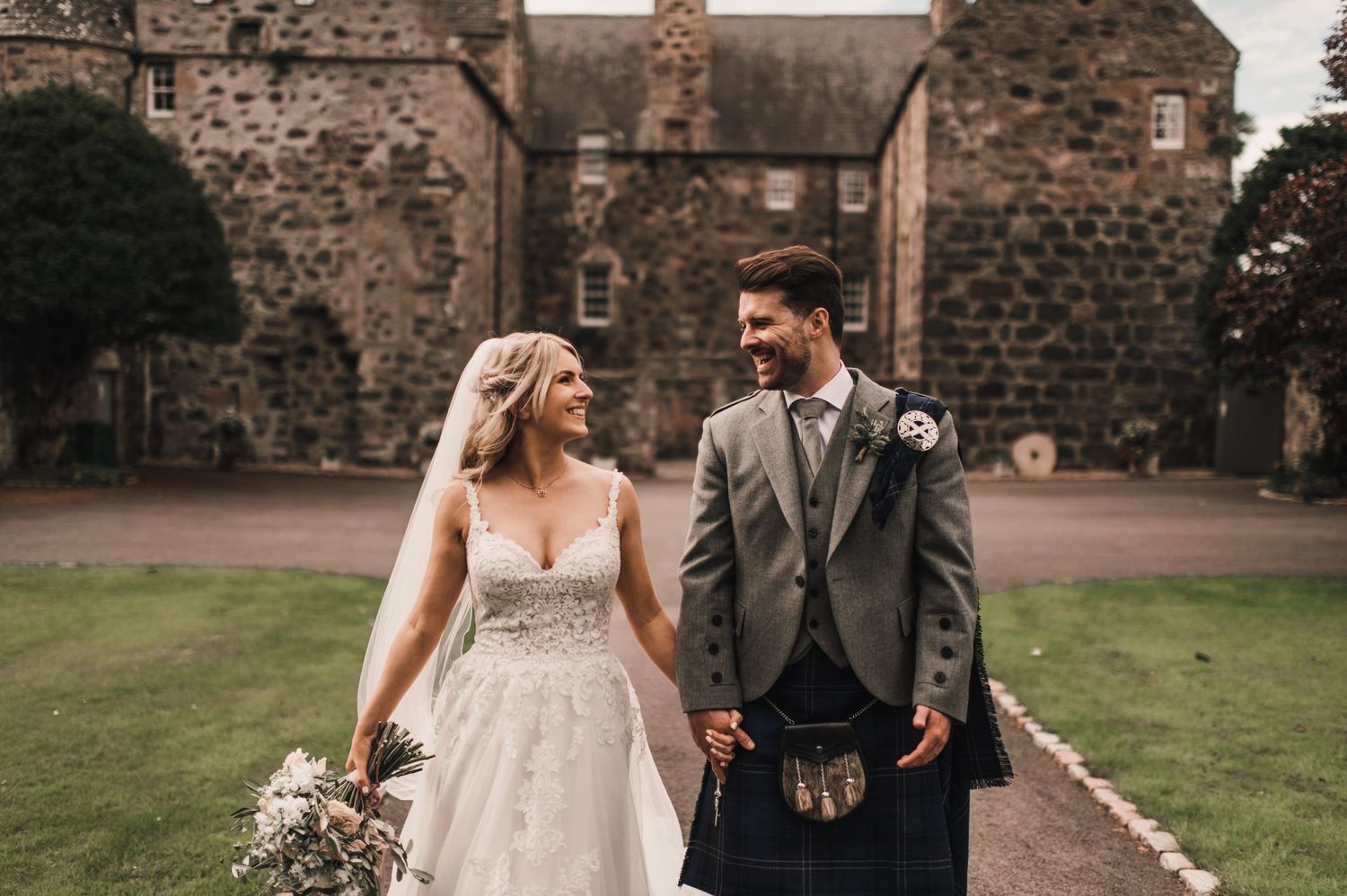 Wedding couple share intimate moment outside historic Scottish stone castle wearing traditional attire.