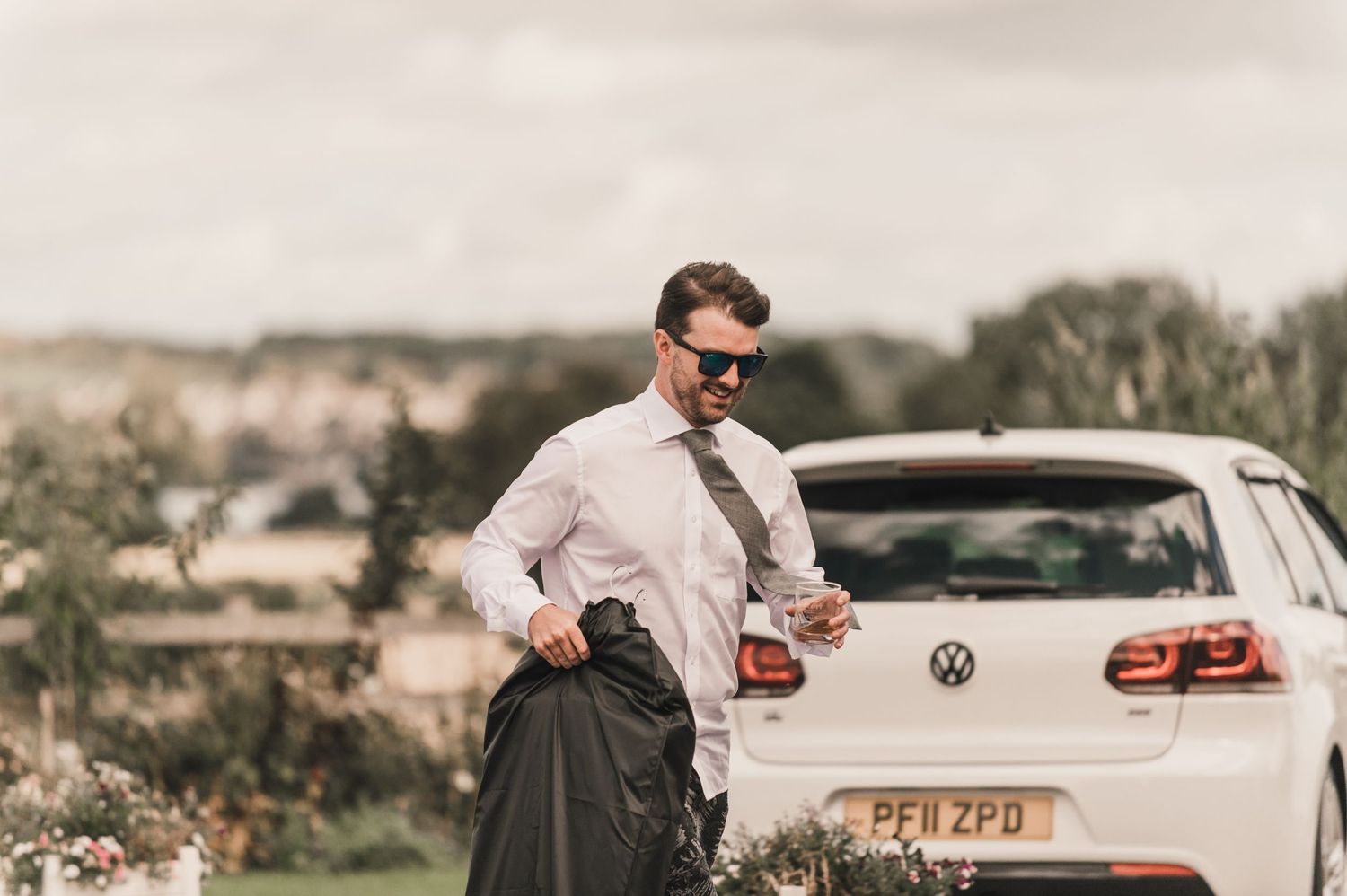 Man in formal attire and kilt stands beside white car in countryside setting.