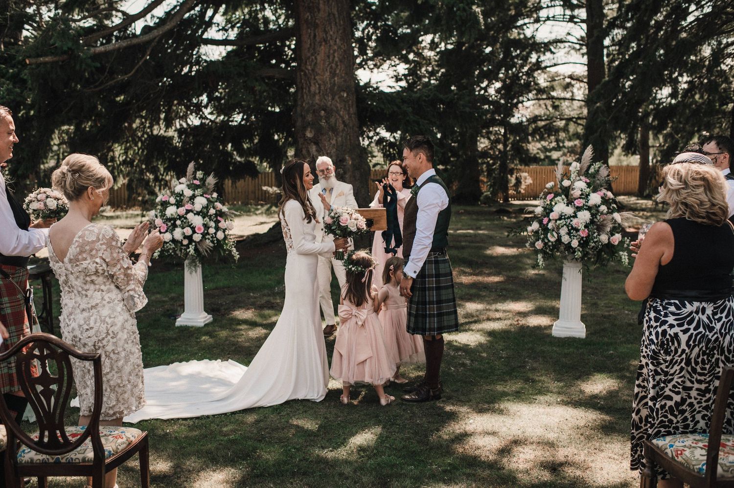 Wedding celebration in a wooded outdoor venue with guests gathered around the ceremony space.